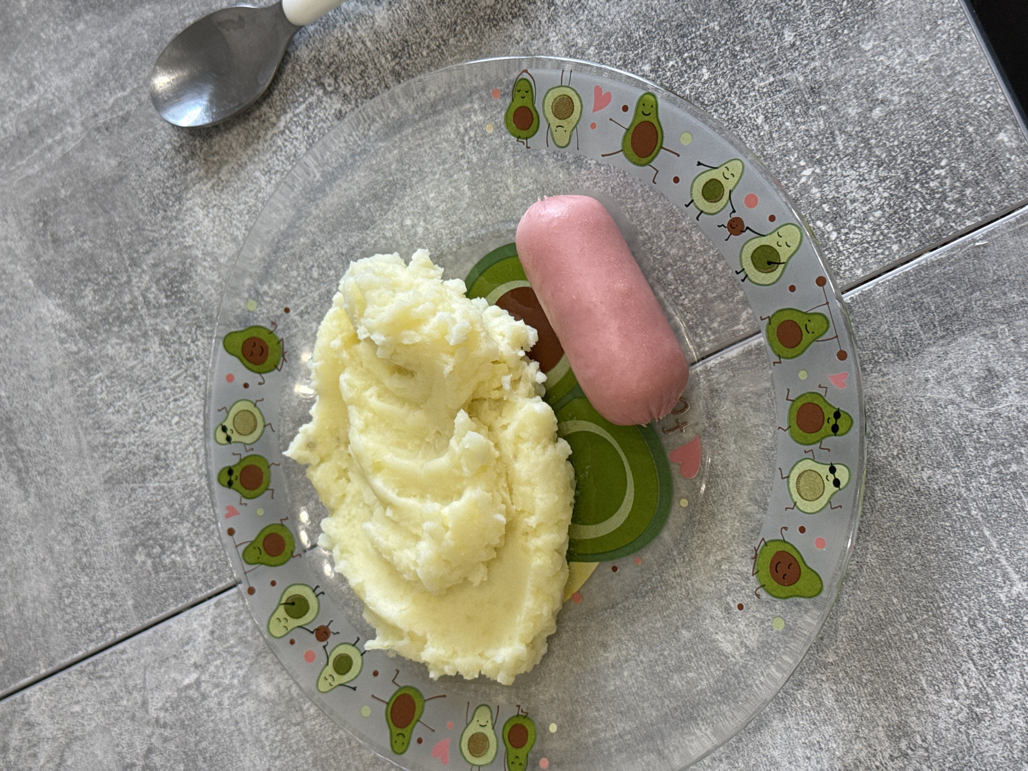 A clear, round plate decorated with cartoon avocados is placed on a grey, textured surface that appears to be a table or countertop made of interlocking panels. On the plate, a serving of mashed potatoes is piled on the left side, and a single pink sausage lies to the right of the potatoes. A silver spoon with a white handle rests to the upper right of the plate. The overall scene suggests a meal setting.
