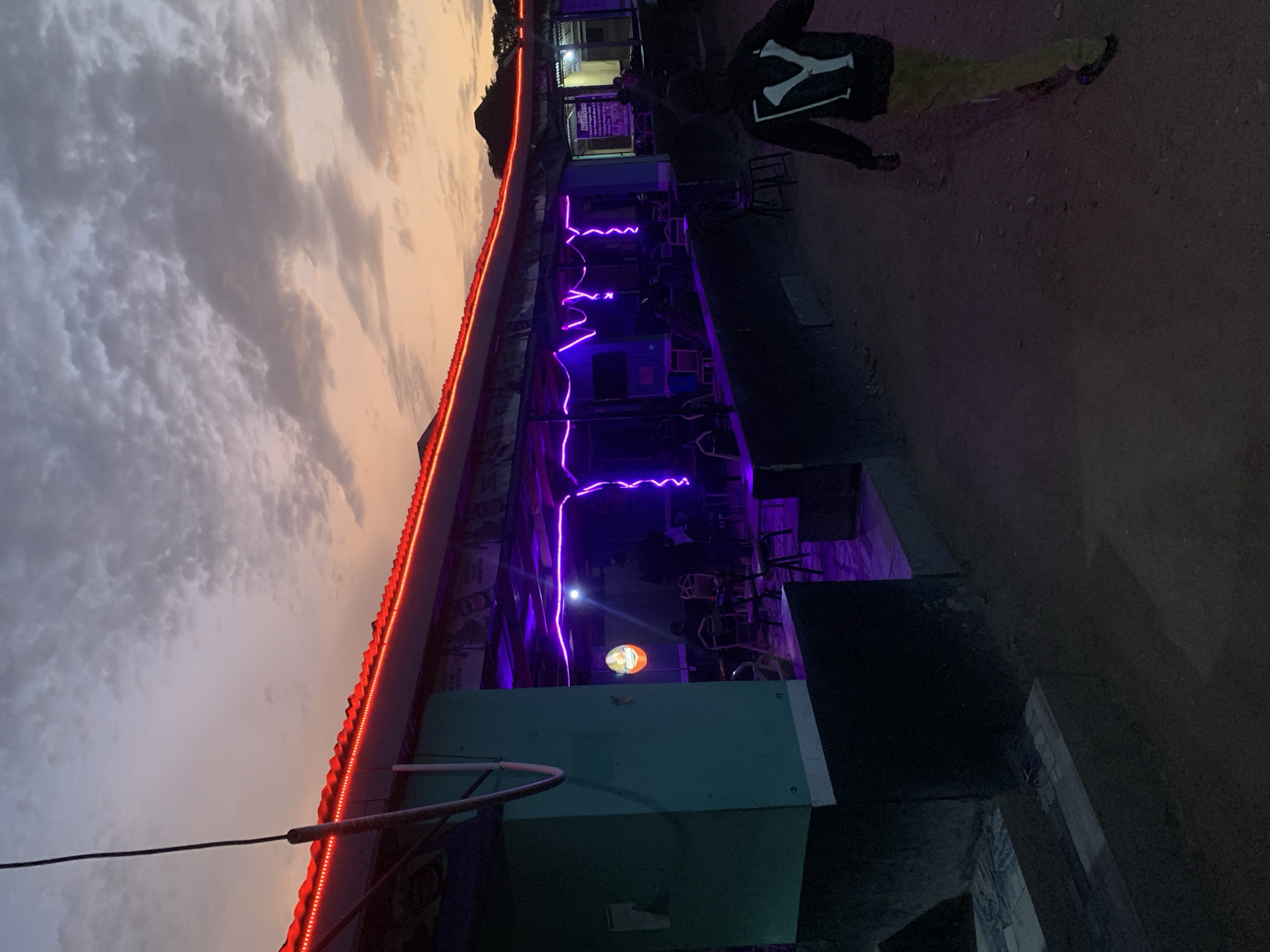 The image depicts an outdoor establishment in Kabong, Nigeria, possibly a bar or restaurant, at dusk. The sky features a mix of cloudy grays and orange hues. The building is adorned with red LED strip lighting along the roofline. Purple neon lights zig-zag below the roofline, illuminating outdoor seating. A figure wearing a black jacket with a white "Y" design, yellow pants, and dark shoes is walking away from the camera in the upper right. A round sign with an illustration is visible. The foreground reveals a green structure, likely a box or container. The ground appears to be a paved or dirt surface.