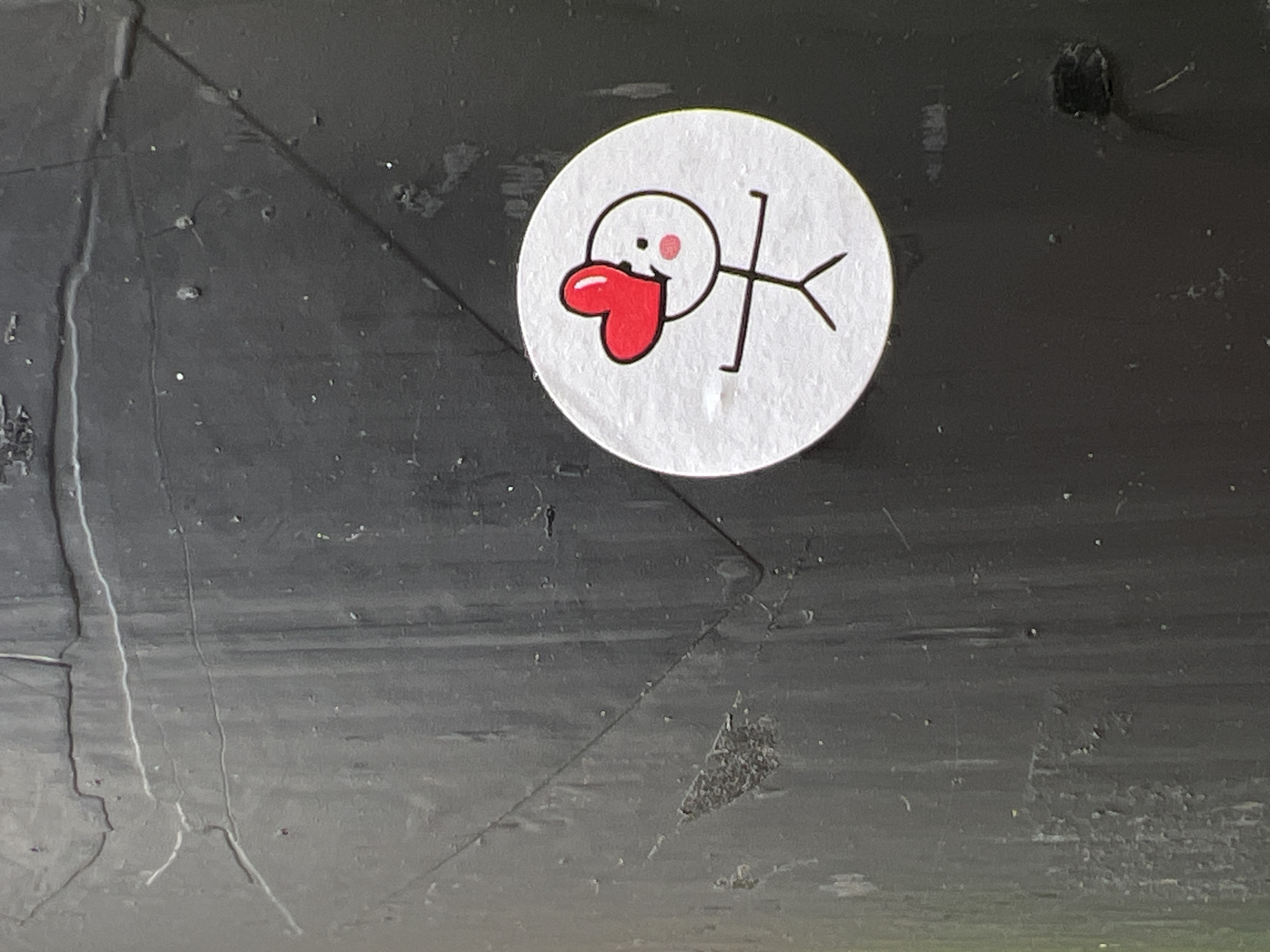 A circular sticker with a cartoon drawing of a stick figure is affixed to a dark, textured, vertical surface. The stick figure has a red heart over its head, two dots for eyes, and two pink circles for cheeks. Its arms are outstretched. The background surface appears to be weathered, with visible lines and a slightly rough texture. The image does not provide any information about its location in Moscow, Russia.