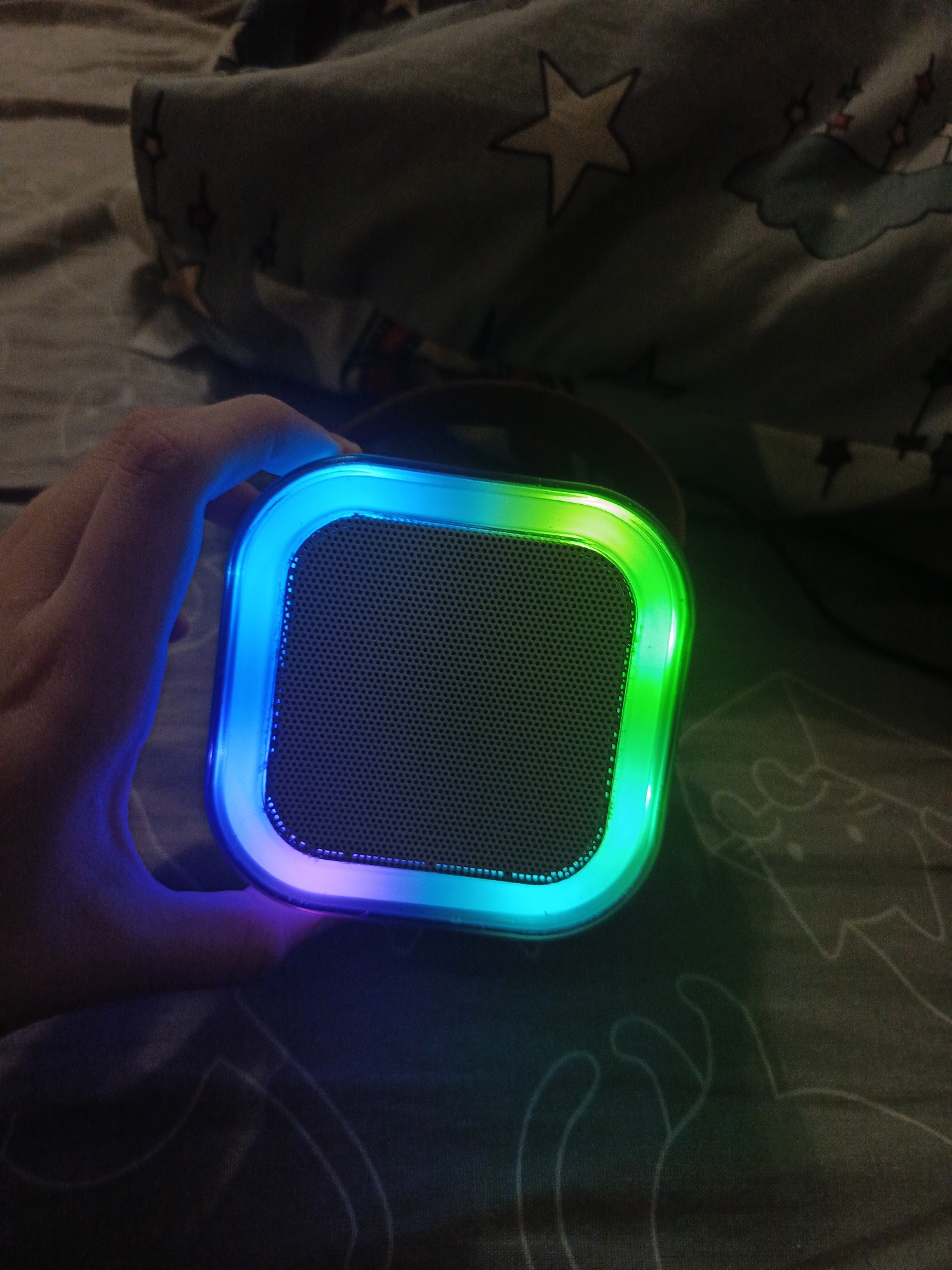 A hand holds a square-shaped wireless speaker with a black mesh grill. The speaker has a light strip around its perimeter that cycles through colors from blue, purple, green and aquamarine. The background consists of a patterned comforter featuring star, moon, and cloud motifs. The location is Unknown, Unknown. No city or further details of location is visible or provided.