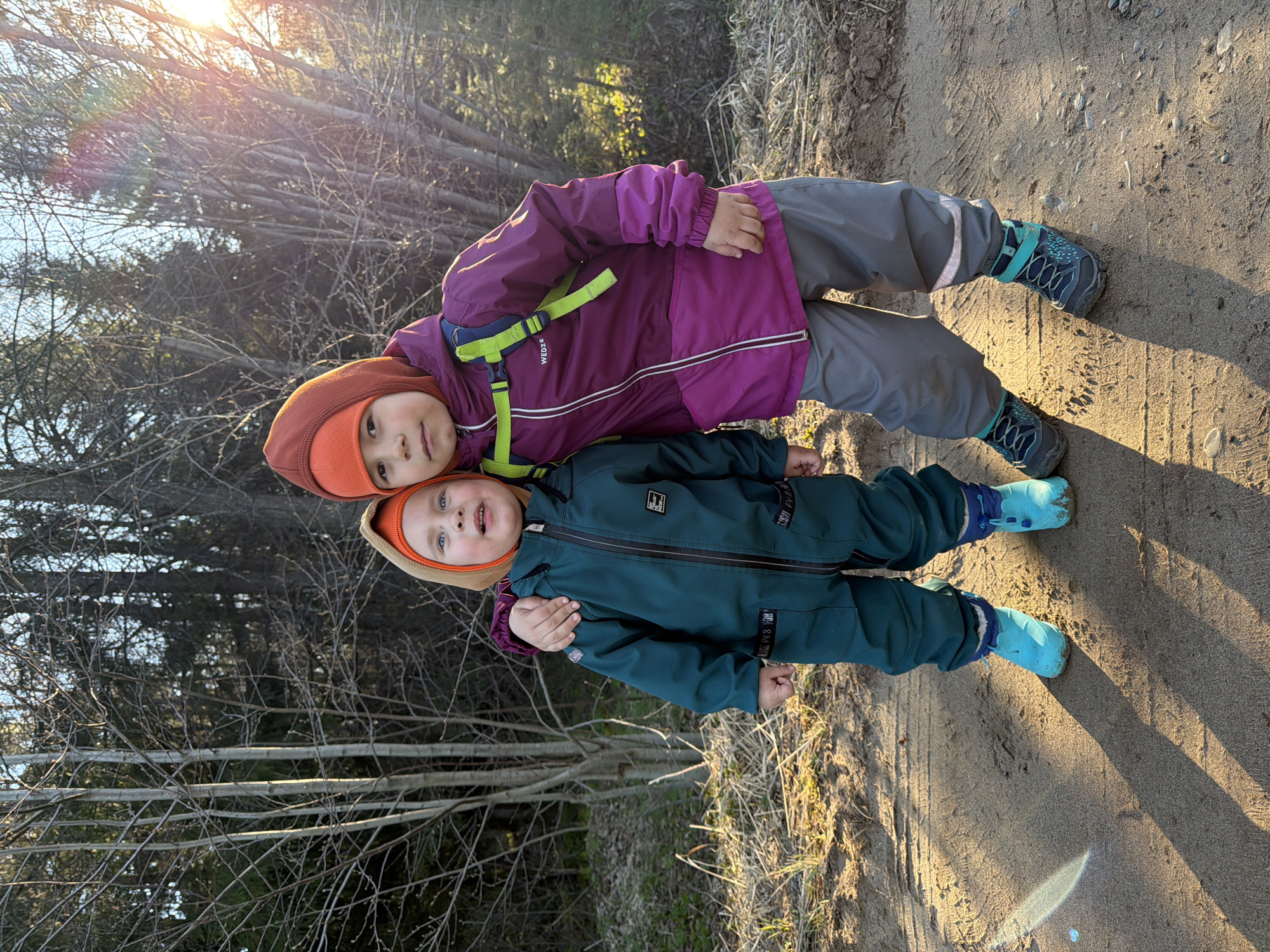 Two children, one with a maroon jacket and grey pants and the other in a green one-piece suit, are standing outside in an environment with trees and sandy soil. The child in the maroon jacket is standing behind the child in the green suit, and their arms are around each other. Both children are wearing orange hats and blue shoes. The setting is in the city of Chaikovsky, Russia. The location appears to be a park or natural area, with trees and sunlight visible in the background.