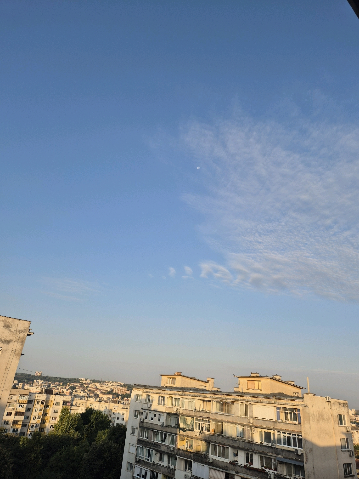 A high-angle shot captures a cityscape bathed in the soft light of early morning or late afternoon. The sky, a clear gradient of pale blue to deeper azure, is adorned with wispy cirrus clouds and a faint, crescent moon. Below, a dense urban landscape unfolds, dominated by numerous apartment buildings of varying heights and architectural styles, many exhibiting signs of age. Trees dot the midground, interspersed with the structures, their dark green foliage providing a contrast to the muted tones of the buildings. In the foreground, the edge of a concrete building, weathered and stained, juts in from the left, its diagonal line leading the eye into the scene. The overall impression is one of a lived-in, everyday urban environment under a tranquil sky.