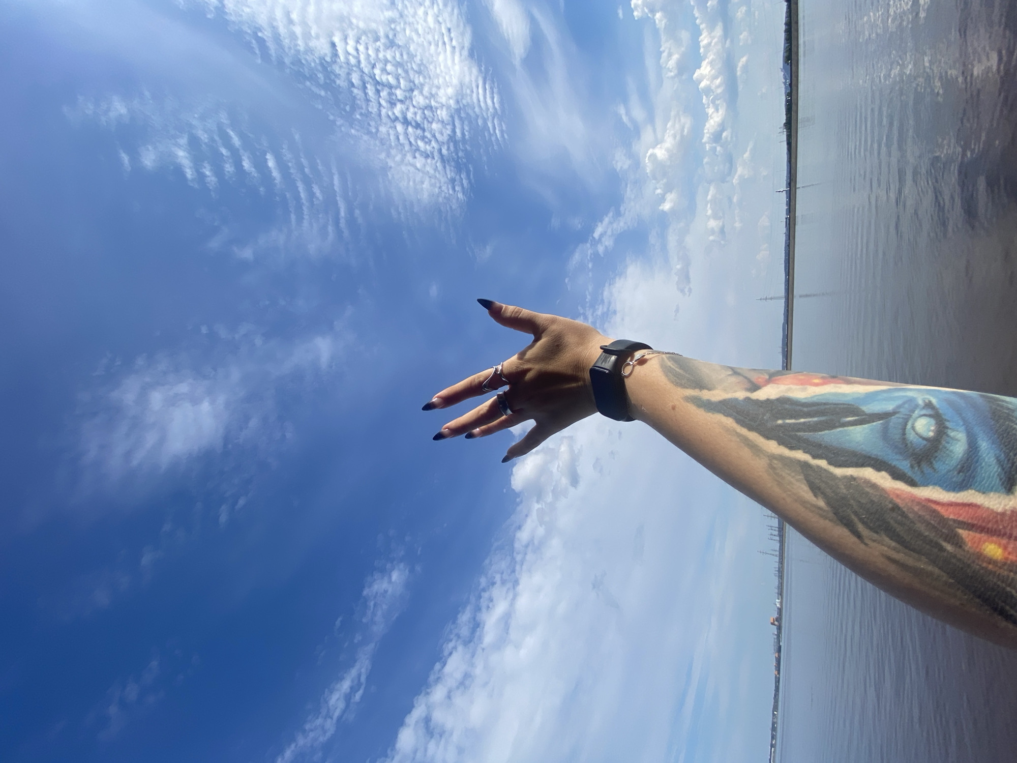 The image features a person's tattooed arm reaching towards a vibrant blue sky filled with wispy white clouds. The arm, adorned with a colorful tattoo of an eye, is extended upwards, with fingers splayed and wearing rings. A black smartwatch is visible on the wrist. The background shows a calm body of water, likely a river or bay, with a distant industrial or urban landscape on the horizon. The scene appears to be taken from a boat or waterfront, with the water occupying the lower portion of the frame. The overall impression is one of freedom and openness, with the hand reaching towards the vast sky. This scene could be located along the Kama River in Chaikovsky, Russia, given the industrial elements and the presence of a large body of water consistent with the river's landscape.