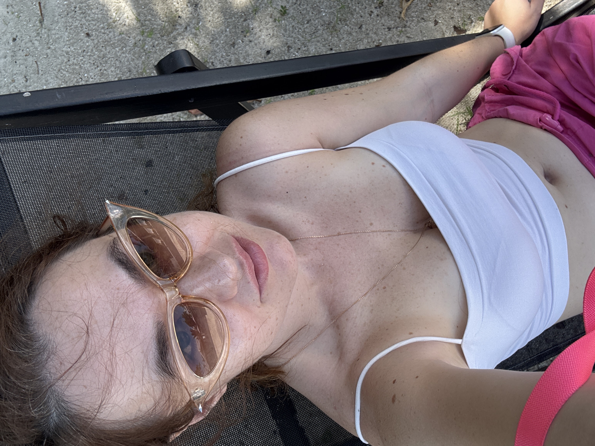 This is a first-person perspective media file of a person relaxing outdoors, likely in Baan Plai Laem, Thailand. The subject is lying on a dark mesh lounge chair, with their body angled towards the camera. They are wearing a white tank top and bright pink shorts, with a pink strap visible across the chest. Light brown sunglasses with a cat-eye frame are worn over the eyes. The person has a delicate gold necklace on, and their skin is lightly freckled.
The background suggests an outdoor setting with concrete or gravel ground and some shadows indicating sunlight, suggesting it is daytime. The lighting appears natural and bright. The overall mood seems relaxed and carefree, as one might feel on a vacation or a leisurely day. The perspective of the camera implies the subject is holding it themselves, creating an intimate and personal view. No visible text is present in the media file.