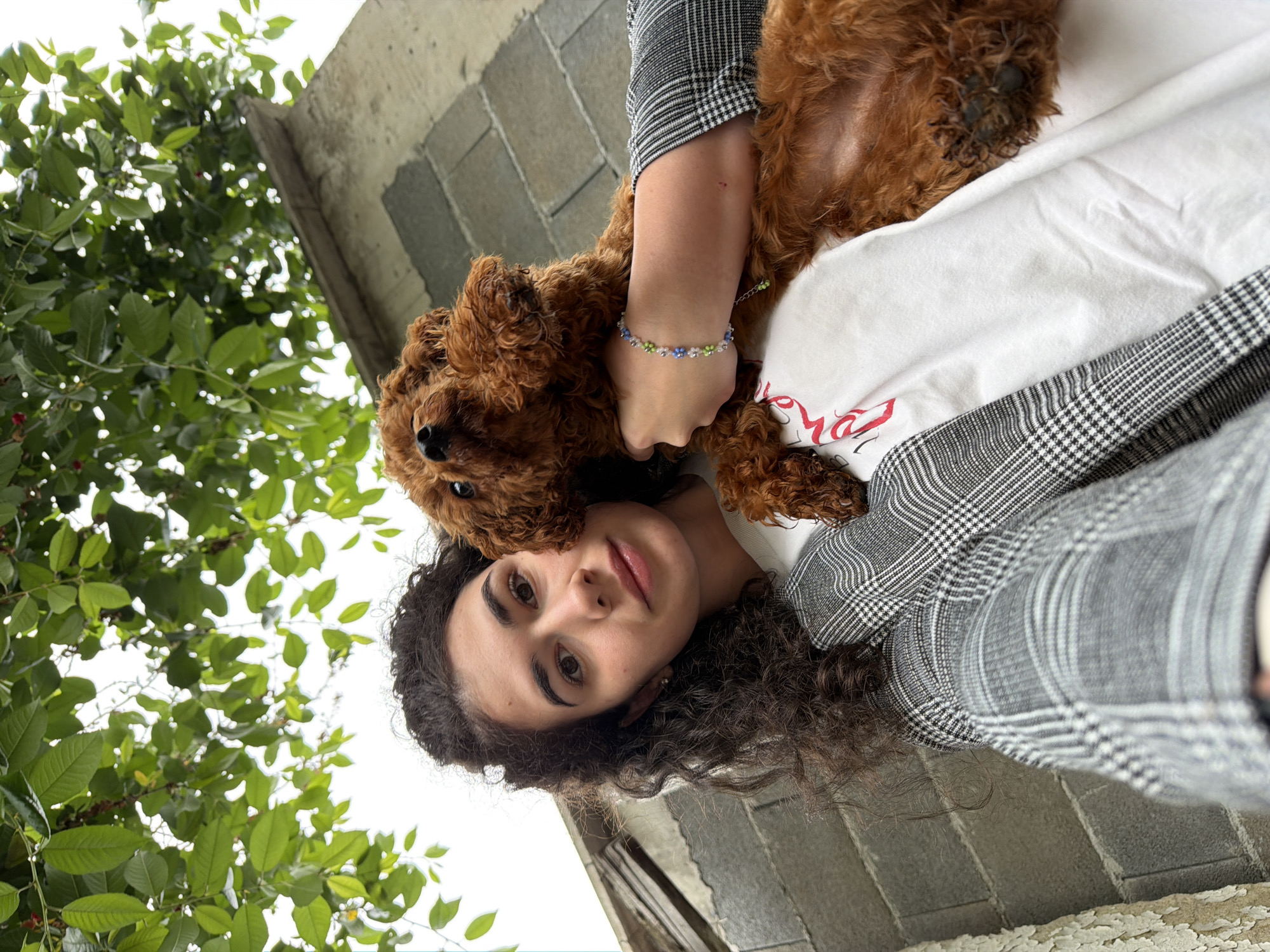 A young woman with curly dark hair is taking a selfie while holding a small, reddish-brown poodle. She is wearing a gray and white checkered blazer over a white t-shirt with red lettering. The dog is nestled against her chest, looking up towards the camera. A bracelet with small colorful beads is visible on the woman's left wrist. The background shows an outdoor setting with green foliage from a tree overhead, a stone wall, and a cloudy sky. The scene appears to be in Sofia, Bulgaria, given the context.