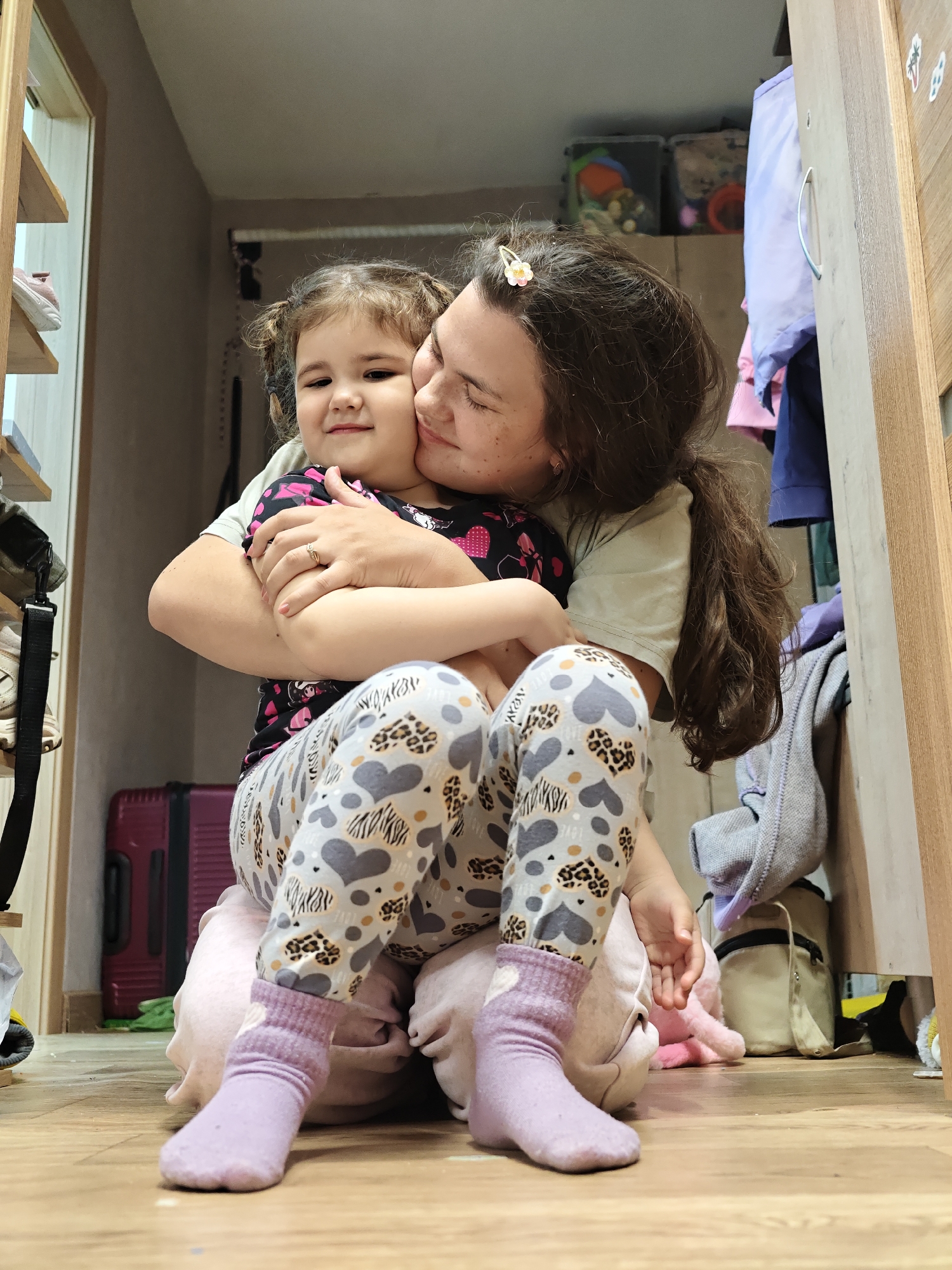 An indoor, low-angle shot shows an adult and a young child sharing a tender moment. The adult, positioned to the right, has long, dark hair with a small white and pink flower clip. She wears a light-colored t-shirt and is embracing the child, pressing her cheek against the child's face as if kissing or nuzzling. Her eyes are closed, conveying affection. A ring is visible on her right hand.
The child, seated between the adult's legs, has dark hair styled in pigtails and looks directly towards the viewer with a slight, sweet smile. She wears a black t-shirt with pink patterns and distinctive leggings featuring grey hearts, leopard print, and zebra stripes. The word "LOVE" is subtly visible within the heart patterns on her leggings. Her feet are clad in light purple socks.
The setting appears to be a cozy, lived-in interior, possibly a closet or small room, with a wooden floor. To the left, a vertical wooden shelving unit with a white shoe is visible. To the right, an open wooden wardrobe reveals various hanging clothes (purples, pinks, blues) and a metal handle on its door. Above the wardrobe opening, two storage bins are visible. A burgundy suitcase is partially seen behind the child on the floor, alongside a beige bag and other items. The scene evokes a feeling of warmth and strong familial bonding, suggesting a moment of comfort and love.