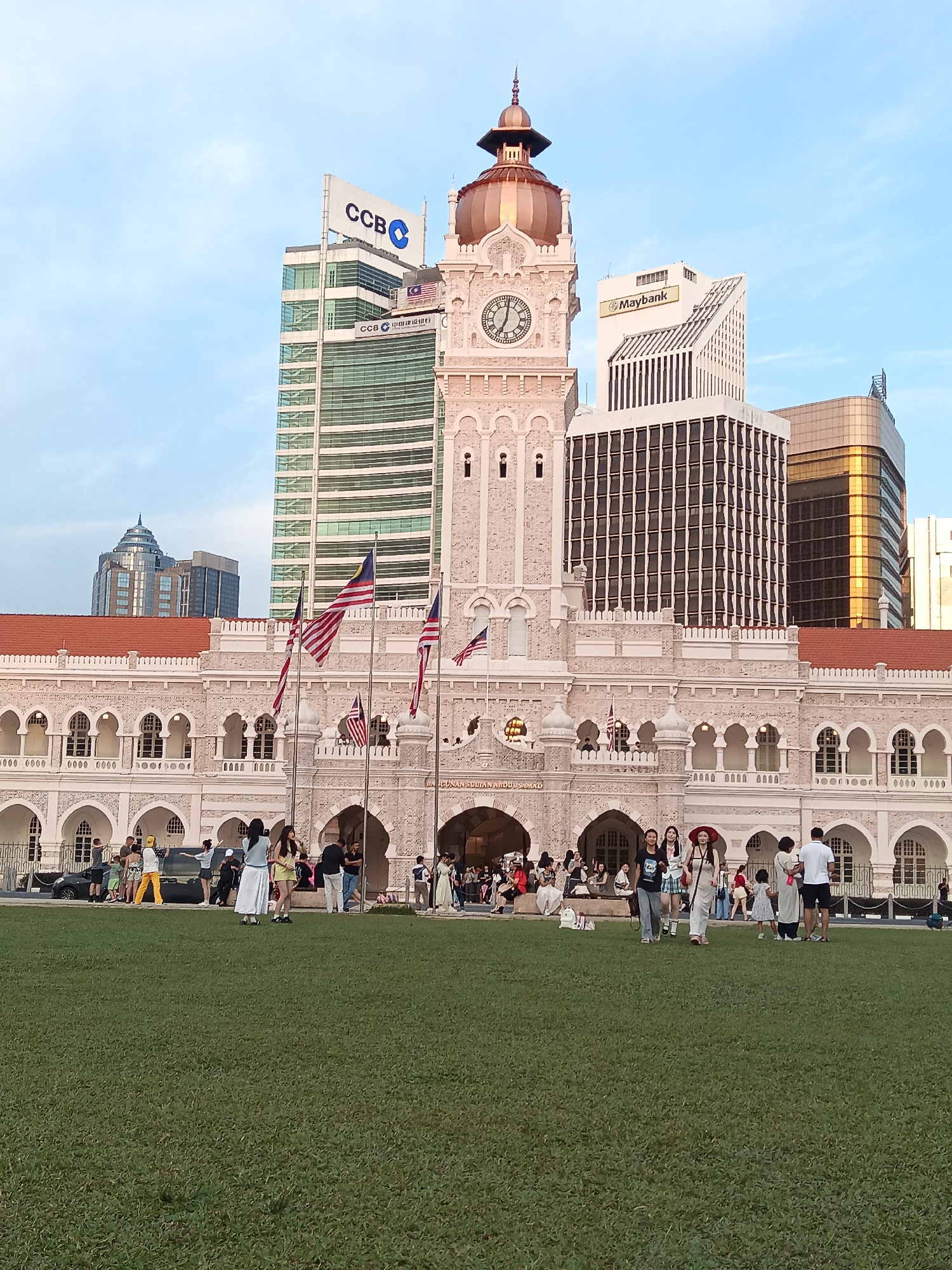 The image shows the Sultan Abdul Samad Building in Kuala Lumpur, Malaysia. The building, a prominent landmark, features colonial architecture with a distinctive clock tower and arched windows. In the foreground, a group of people are gathered on a manicured green lawn, some walking and others standing. Behind the building, modern skyscrapers rise into the sky, one of which displays the "Maybank" logo, and another a "CCB" logo. The Malaysian flag is visible flying on poles in front of the building. The sky is a clear blue with some light clouds, indicating it is daytime, likely late afternoon given the lighting.