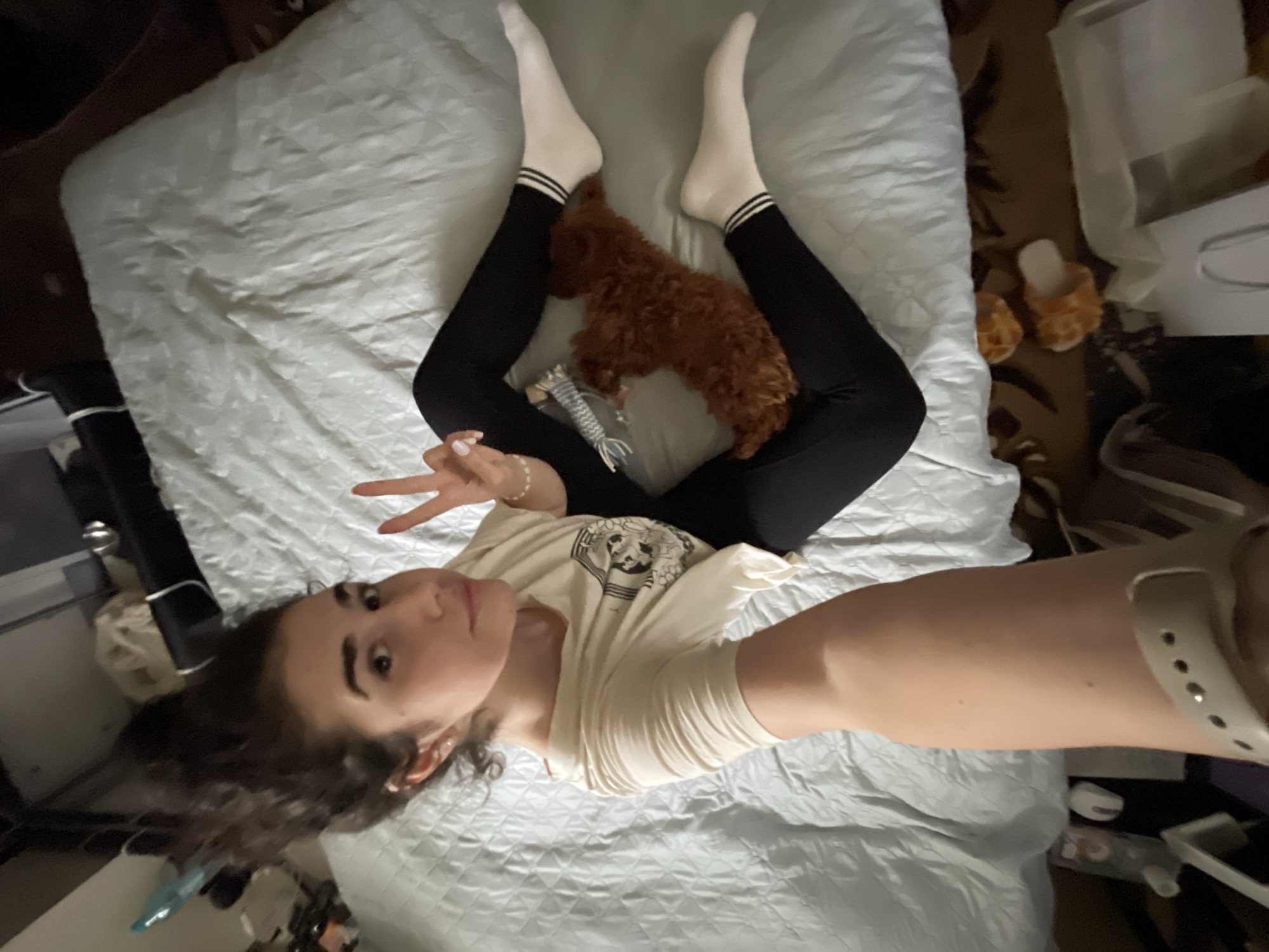 An overhead selfie captures a young woman lying on a bed, looking towards the camera, making a peace sign with her left hand. She has dark, curly hair and is wearing a light-colored t-shirt with an unidentifiable circular graphic design, black leggings, and white ankle socks with two black stripes near the cuffs. A small, reddish-brown dog, possibly a poodle or doodle mix, is curled up asleep between her legs, near a striped, rope-like dog toy.
The bed is covered with a light gray or white quilted comforter. To the woman's right, on the floor beside the bed, are a pair of fluffy, orange-toned slippers and a white paper bag or box, along with other indistinct items. Her right arm extends towards the camera, and a light-colored smartwatch or fitness tracker is visible on her right wrist. The setting appears to be indoors, likely a bedroom, with the overall ambiance suggesting a relaxed moment. No specific time of day or weather is discernible from the indoor environment. The location context for this scene is Sofia, Bulgaria. No legible text is visible within the image other than the graphic on her t-shirt, which is too indistinct to decipher.