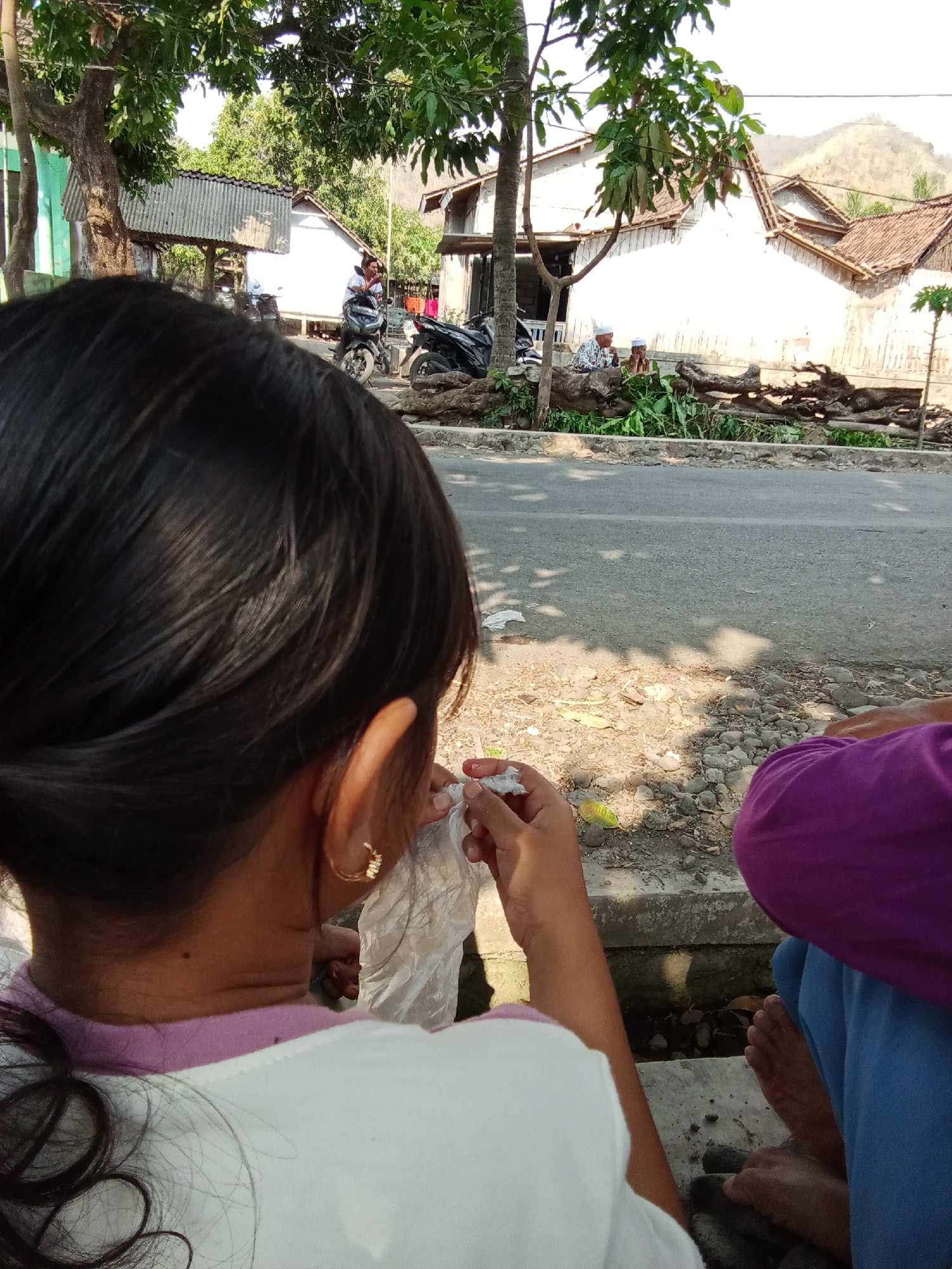 In Trebungan, Indonesia, a person with dark hair, wearing a white shirt with a pink collar and a gold hoop earring, is sitting by the side of a paved road. Their back is to the camera, and they are holding a white plastic object in their hands. To the right, another person is visible, wearing a purple top and blue pants, with their legs and feet exposed. They are sitting on a concrete ledge. The background shows several white buildings with tiled roofs, some trees, and a motorcycle parked in front of one of the buildings. A person is seated on the motorcycle, wearing a white shirt. In the distance, a hillside is visible. The ground is covered in gravel and dirt, with some foliage and tree branches scattered around. The scene appears to be a daytime outdoor setting.