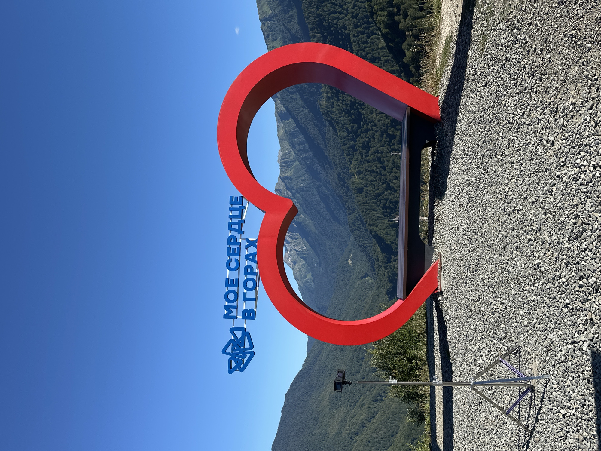 The image shows a large, red, heart-shaped bench with a wooden seat, positioned on a gravel surface. Above the heart, blue text reads "МОЕ СЕРДЦЕ В ГОРАХ" which translates to "MY HEART IN THE MOUNTAINS". To the left of the bench, a camera on a tripod is set up. The background features a lush, green mountain range under a clear blue sky. This monument is a popular attraction located in the mountain resort of Estosadok, in Russia.