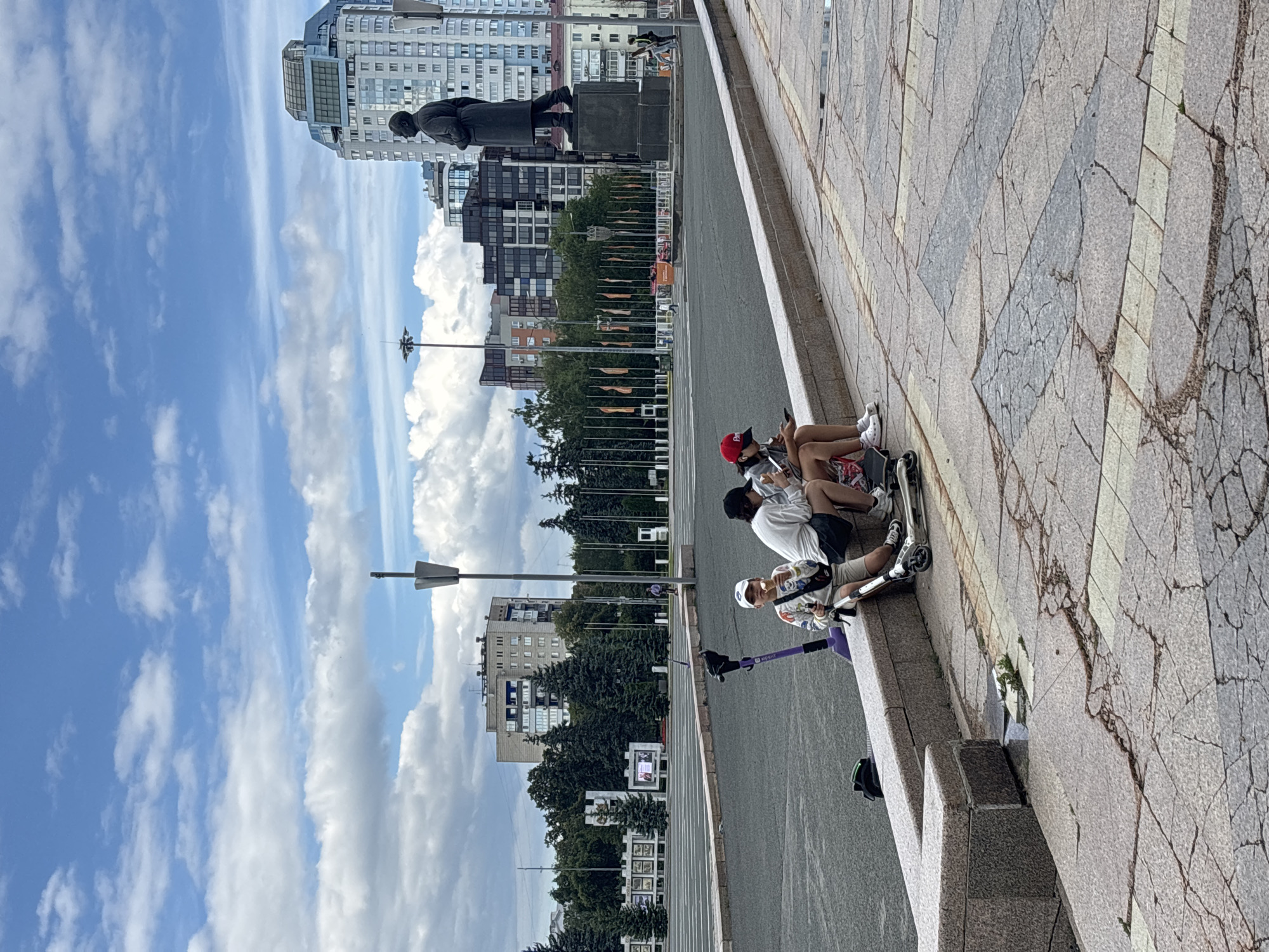 The image depicts a wide, open urban plaza in Novokuybyshevsk, Russia. The foreground features a low, granite retaining wall where three young individuals are seated, engrossed in their mobile devices and enjoying ice cream. Two scooters are positioned near them, one purple and one silver. The plaza floor is paved with large, cracked stone tiles, some with distinct geometric patterns.
In the middle ground, a vast, empty asphalt area stretches towards a line of tall evergreen trees. Beyond the trees, several apartment buildings of varying architectural styles are visible, with a prominent, modern skyscraper dominating the right side of the frame. To the left of the skyscraper, a large, dark statue on a pedestal stands silhouetted against the sky. Flags are displayed along a central avenue leading towards the statue.
The sky is a bright blue, dotted with scattered white cumulus clouds. The overall impression is one of a quiet, sunny day in a public urban space.