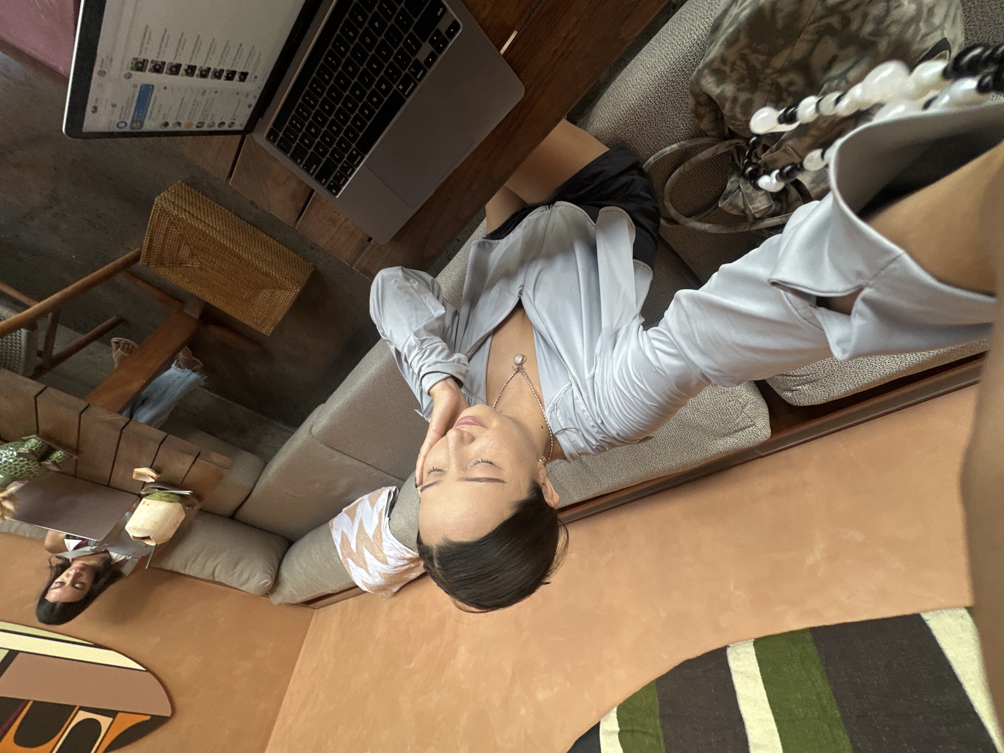 The image depicts an indoor cafe scene in North Kuta, Indonesia. The primary subject is a woman sitting on a sofa, facing a laptop placed on a wooden table. She is wearing a light blue zip-up jacket, a black skirt, and a necklace with a pendant. Her head is tilted down, and her right hand is resting on her cheek, suggesting contemplation or fatigue.
To the right of the primary subject, another woman is seated at a table, also looking at a laptop. A whole coconut with a straw is placed on the table in front of her.
The environment includes a long L-shaped sofa, wooden tables, and walls decorated with artwork. A woven basket is visible on the floor beneath a table. The overall impression is that of a relaxed, work-conducive setting, possibly a cafe or co-working space.