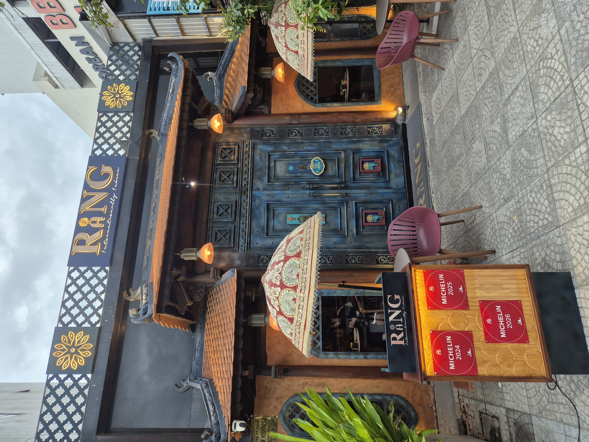 The image shows the exterior of RANG, an internationally Indian restaurant located in Đà Nẵng, Vietnam. The restaurant features a distinctive architectural style with a dark facade, a tiled roof with curved eaves, and a large, ornate blue door. Two decorative umbrellas with intricate patterns are placed outside the entrance. A display stand prominently shows three red plaques, each with "MICHELIN" and a year: 2024, 2025, and 2026. Two purple chairs and a table are visible on the tiled sidewalk in front of the restaurant. The overall scene suggests a well-established establishment with Michelin recognition.
