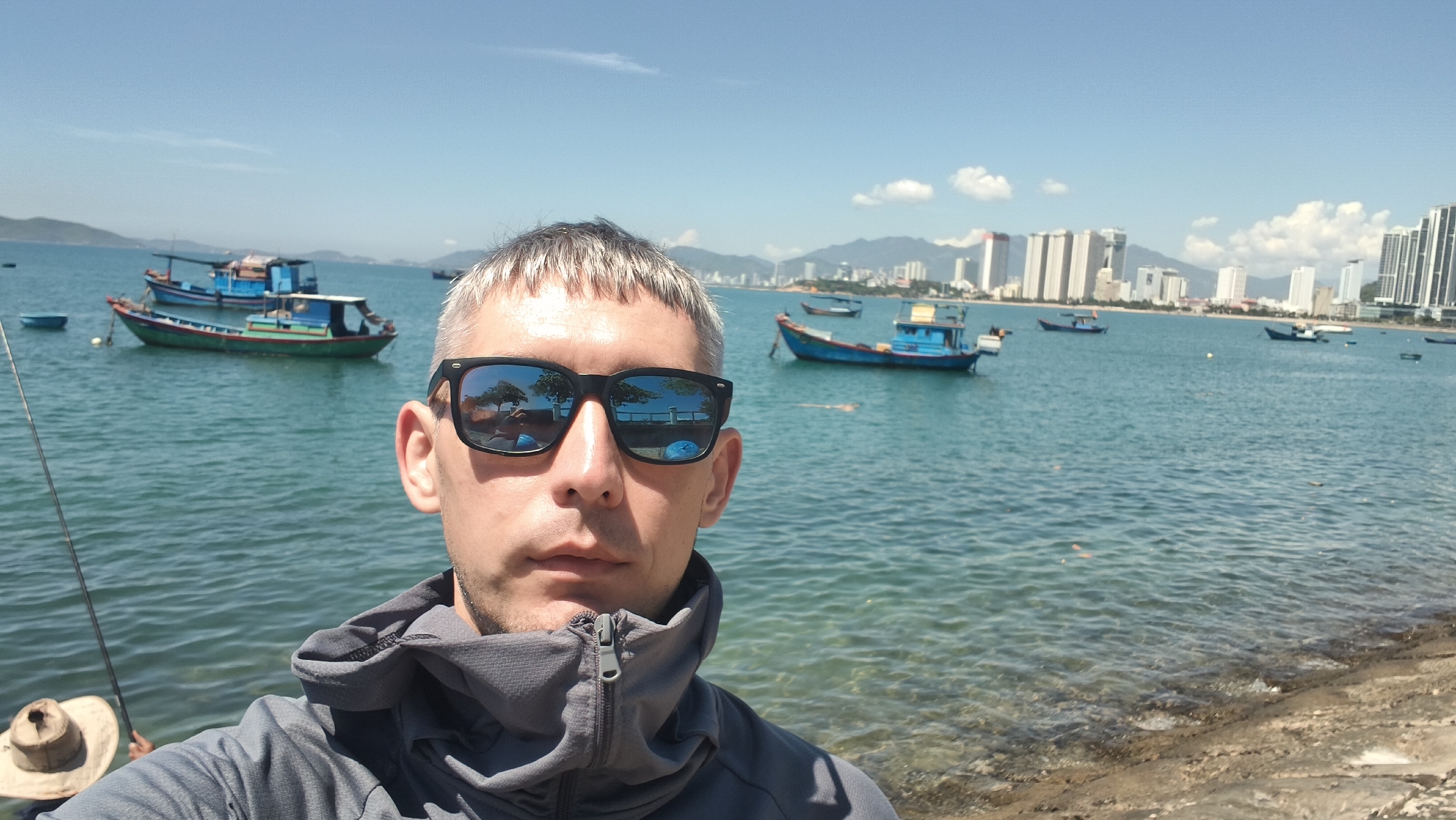 A man wearing sunglasses and a grey hooded jacket is taking a selfie by the sea. In the background, several boats are anchored in the water. A city skyline with tall buildings is visible in the distance. The man's sunglasses reflect the scene in front of him, showing trees and a waterfront promenade. The image appears to be taken in Bac Nha Trang Ward, Vietnam, given the presence of the city skyline and the style of boats. The sea is calm and the sky is clear with some clouds. A fishing rod is visible to the left of the man.
