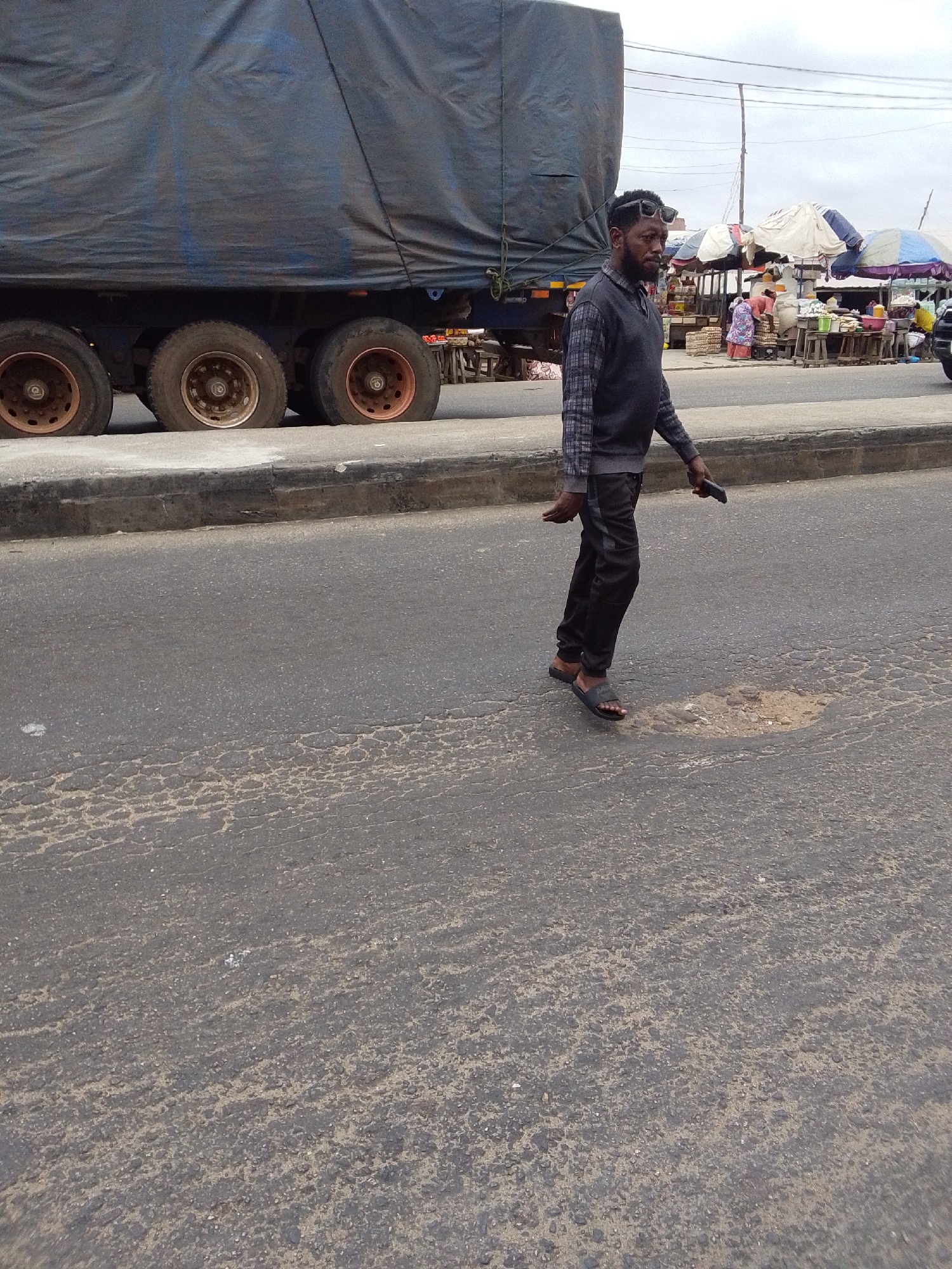 This daylight image depicts a bustling urban street scene in Lokoja, Nigeria, under an overcast sky. The main subject is a dark-skinned man walking from right to left across an asphalt road. He is dressed in a long-sleeved plaid shirt layered with a dark sleeveless vest, dark trousers, and black slides. Sunglasses are perched on his head, and he sports a beard. He holds a dark-colored smartphone in his right hand.
To the left, a large truck or trailer is parked or moving slowly, its cargo covered by a dark blue tarp. Its multiple wheels, featuring visible rust on the rims, are positioned alongside a concrete curb that separates the road from a narrow verge. The road surface is visibly worn, characterized by cracks and a distinct pothole near the man's feet.
In the background, beyond the truck, a vibrant market area is active. Numerous stalls, shaded by various white, blue, and striped tarpaulin umbrellas, are set up. They display a range of goods on wooden tables and stands. Several people, likely vendors and shoppers, are visible amidst the market activity. Utility poles can be seen against the cloudy sky in the distance. No discernible text is visible in the image.