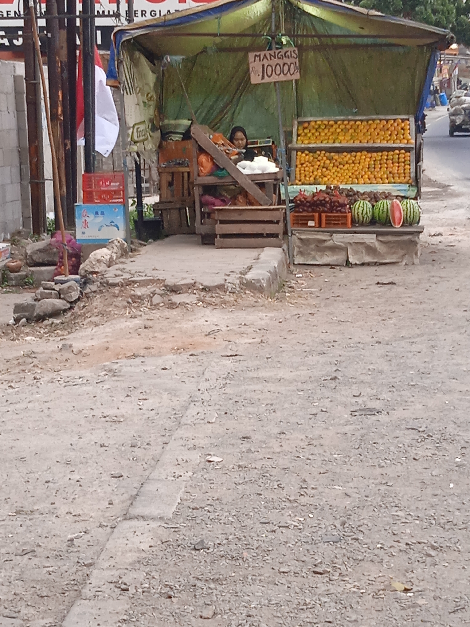 The image depicts an outdoor fruit stand in Cileunyi, Indonesia, by a roadside. The stand, covered by a green tarp, displays various fruits. Oranges are prominently featured in two stacked wooden crates behind a woman seated at the stand. In front of the oranges, a selection of red fruits, possibly berries or small tomatoes, are displayed in orange plastic crates. To the right, two whole watermelons and one half-cut watermelon sit on a stone surface. A wooden sign with the text "MANGGIS Rp. 10000" is visible near the oranges, indicating mangosteens are for sale at 10,000 Rupiah. The ground surrounding the stand is a mixture of dirt and gravel. In the background, a road can be seen with a vehicle passing by. Several poles, one of which has a red and white Indonesian flag attached, are to the left of the stand. Various items, including red crates and a blue box with Asian characters, are scattered near the base of the poles.