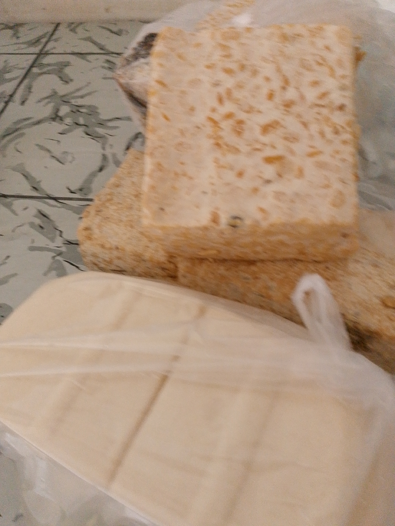 The image shows a close-up of a block of tempeh, a traditional Indonesian soy product, placed on a wooden cutting board. The tempeh is light brown with visible soy beans. Underneath the cutting board, a stack of flat, pale yellow dough or pastries is visible, wrapped in clear plastic. The background is a tiled surface with a green and gray marbled pattern. The scene appears to be in a domestic setting, possibly a kitchen. The lighting is soft, creating some shadows. No people are visible in the image. The location is specified as Babadan, Indonesia.