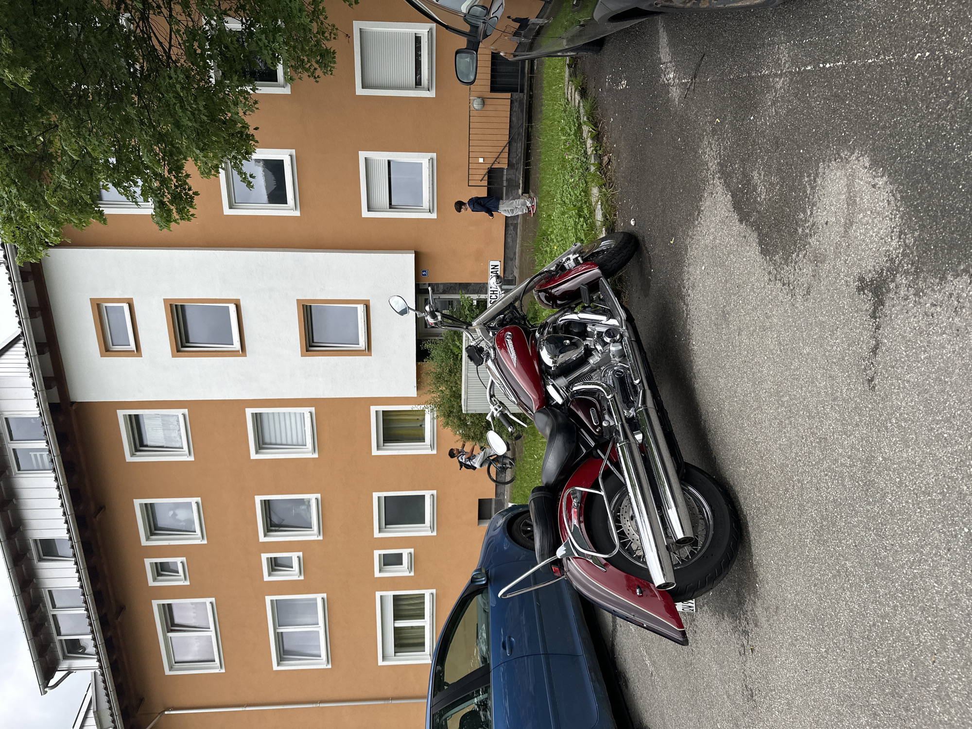 This image shows a street scene in Garmisch-Partenkirchen, Germany. In the foreground, a red motorcycle is parked on asphalt. To the left, a blue car is parked. Behind the motorcycle and cars, there is a residential building with orange and white facade. Several windows are visible on the building. A young boy is riding a bicycle on the grass area in front of the building. Another young boy is standing on the grass area. A tree with green leaves is partially visible on the right side of the image. The weather appears overcast.