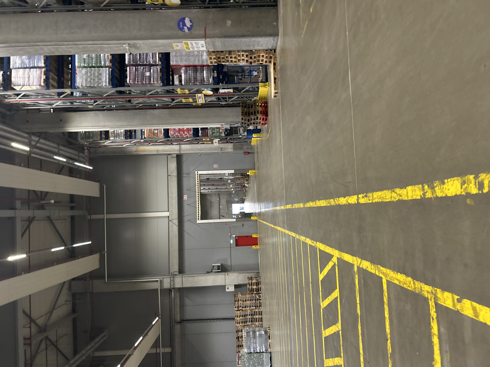 The image shows the interior of a large warehouse in Stryama, Bulgaria. The warehouse features high ceilings with industrial lighting and exposed concrete pillars. Tall shelving units filled with pallets of goods, likely beverages or food items, dominate the right side of the frame. The floor is concrete, marked with yellow lines indicating walkways or parking spaces. In the mid-ground, a red door is visible on a grey wall. Pallets of stacked items are also present in various areas of the warehouse. The overall impression is one of a functional, industrial storage facility.
