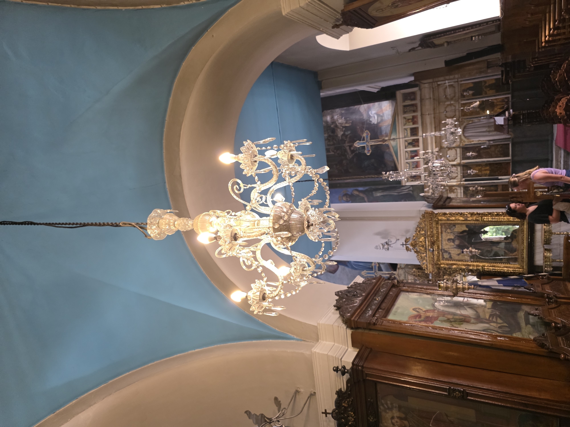 The image shows the interior of a church in Chania, Greece. A large, ornate crystal chandelier hangs from the ceiling, illuminating the vaulted blue ceiling and surrounding arches. The church features religious iconography, including large framed paintings and icons displayed in gilded frames. Two women are present, standing near the center of the frame, conversing. One woman, on the left, wears a black top and sunglasses on her head. The other woman, on the right, wears a purple and white striped tank top. The overall scene depicts the solemn and ornate atmosphere typical of a historic religious establishment.