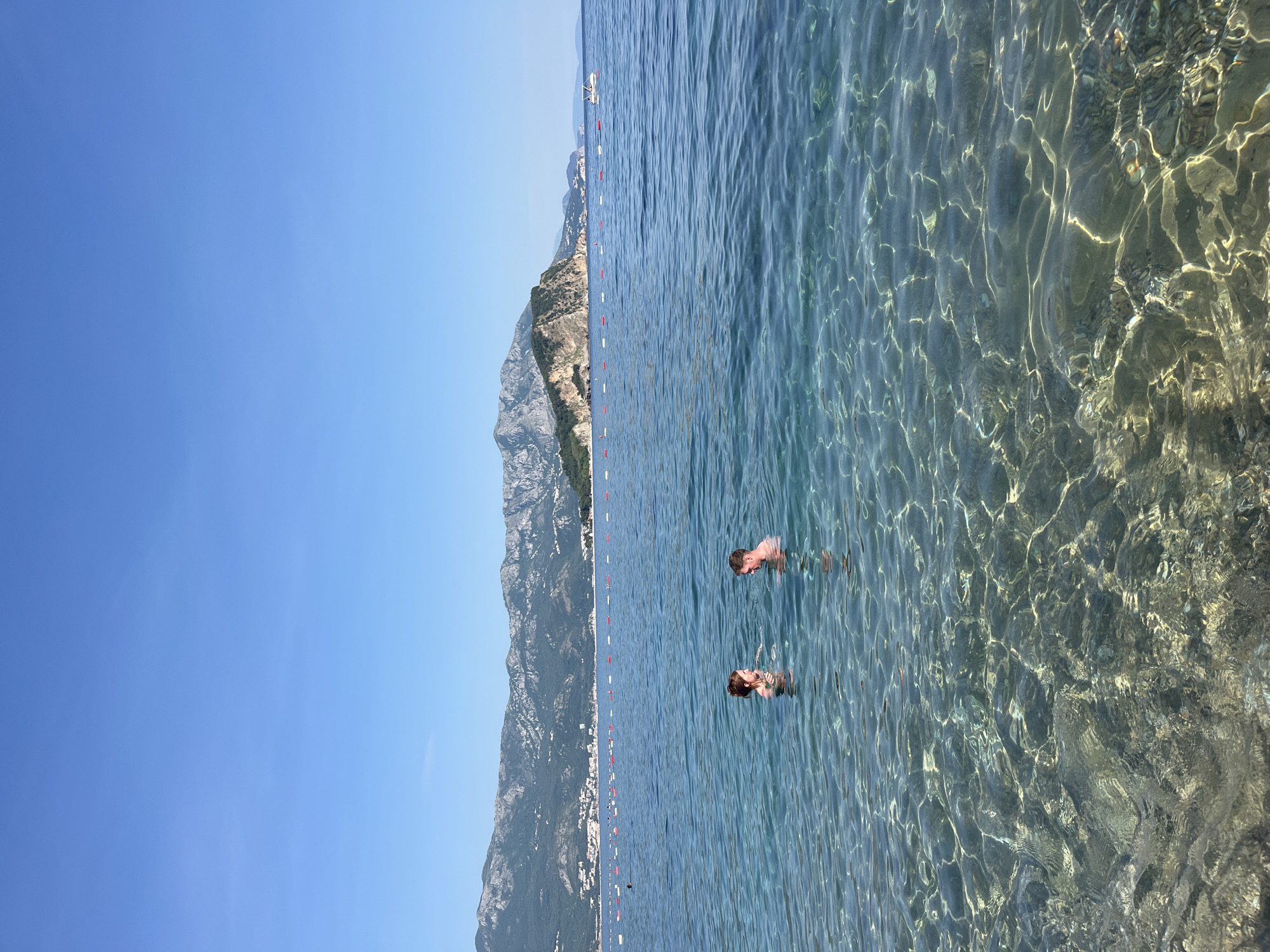 A serene coastal scene is captured in a vertically oriented, outdoor photograph. The image prominently features a clear, sun-dappled body of water where two individuals are swimming. The water's transparency reveals the sandy bottom and underwater textures.
In the background, a mountainous coastline stretches along the horizon, providing a stark contrast to the clear blue sky. The mountains are covered with dense greenery and rocky outcrops, giving a natural, rugged feel to the scenery.
The scene is captured in the city of Budva, Montenegro. The location shows the natural beauty of the coastline and the clear waters of the Adriatic Sea.