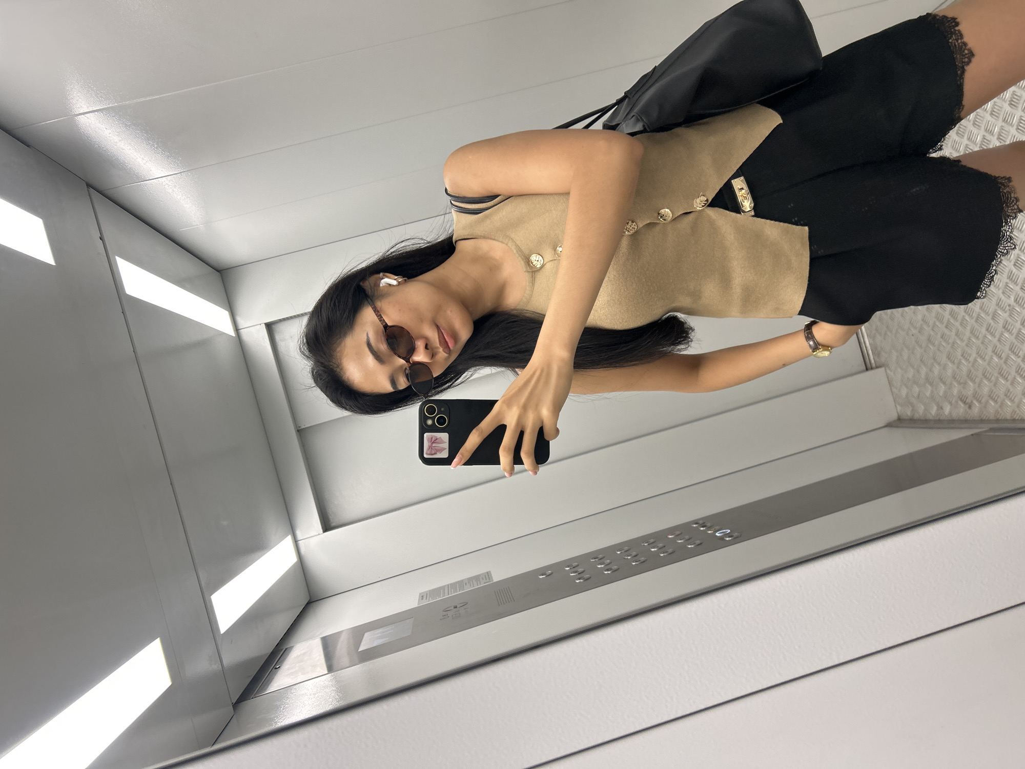 A young Asian woman is taking a mirror selfie in an elevator. She is wearing a beige sleeveless top with gold buttons, black shorts with lace trim, sunglasses, and white wireless earbuds. She is holding a black smartphone with a pink bow sticker on the back. The elevator has gray walls and a metal control panel with buttons. The floor is metal with a diamond plate pattern. The scene appears to be in Moscow, Russia.