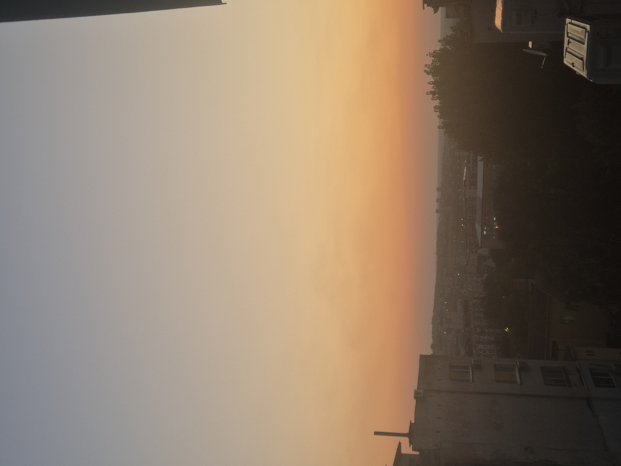 The image captures a cityscape at dusk or dawn. The sky transitions from a pale blue at the top to a warm orange and yellow hue near the horizon, indicating the presence of the sun below the frame. In the foreground, a multi-story building with a rough, textured facade is visible on the left. The building has several windows, and what appears to be an air conditioning unit mounted on its exterior. To the right and in the mid-ground, a dense cluster of trees obscures a portion of the city. The background reveals a sprawling urban landscape with numerous buildings, some with visible lights, suggesting a residential or commercial area. The overall atmosphere is tranquil and serene, characteristic of twilight. The exact location is not identifiable from the image.