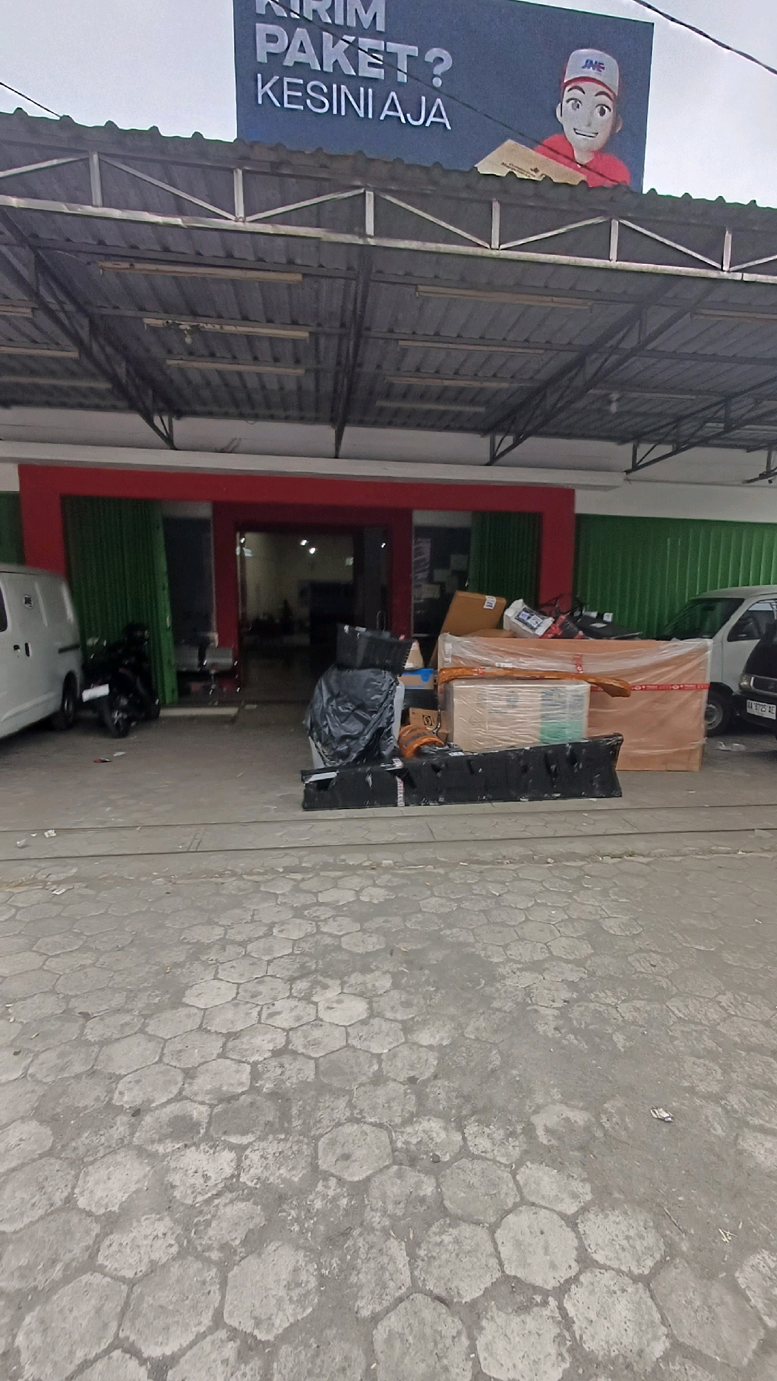 The image captures the exterior of a logistics or delivery service building in Kedu, Indonesia, possibly a JNE or similar service. Above the building, a large banner displays text in Indonesian, including "KIRIM PAKET?" and "KESINI AJA," translating to "SEND A PACKAGE?" and "COME HERE." A cartoon illustration of a delivery person holding a package is also on the banner. The building itself has a red-trimmed entrance with green roll-up doors. Outside the entrance, several packages are piled up, some wrapped in plastic and cardboard boxes. A white van is parked to the left of the entrance, and a black motorcycle is parked near the van. The ground in front of the building is paved with hexagonal concrete blocks. The overall scene suggests a busy delivery hub or a location where packages are being stored and dispatched.