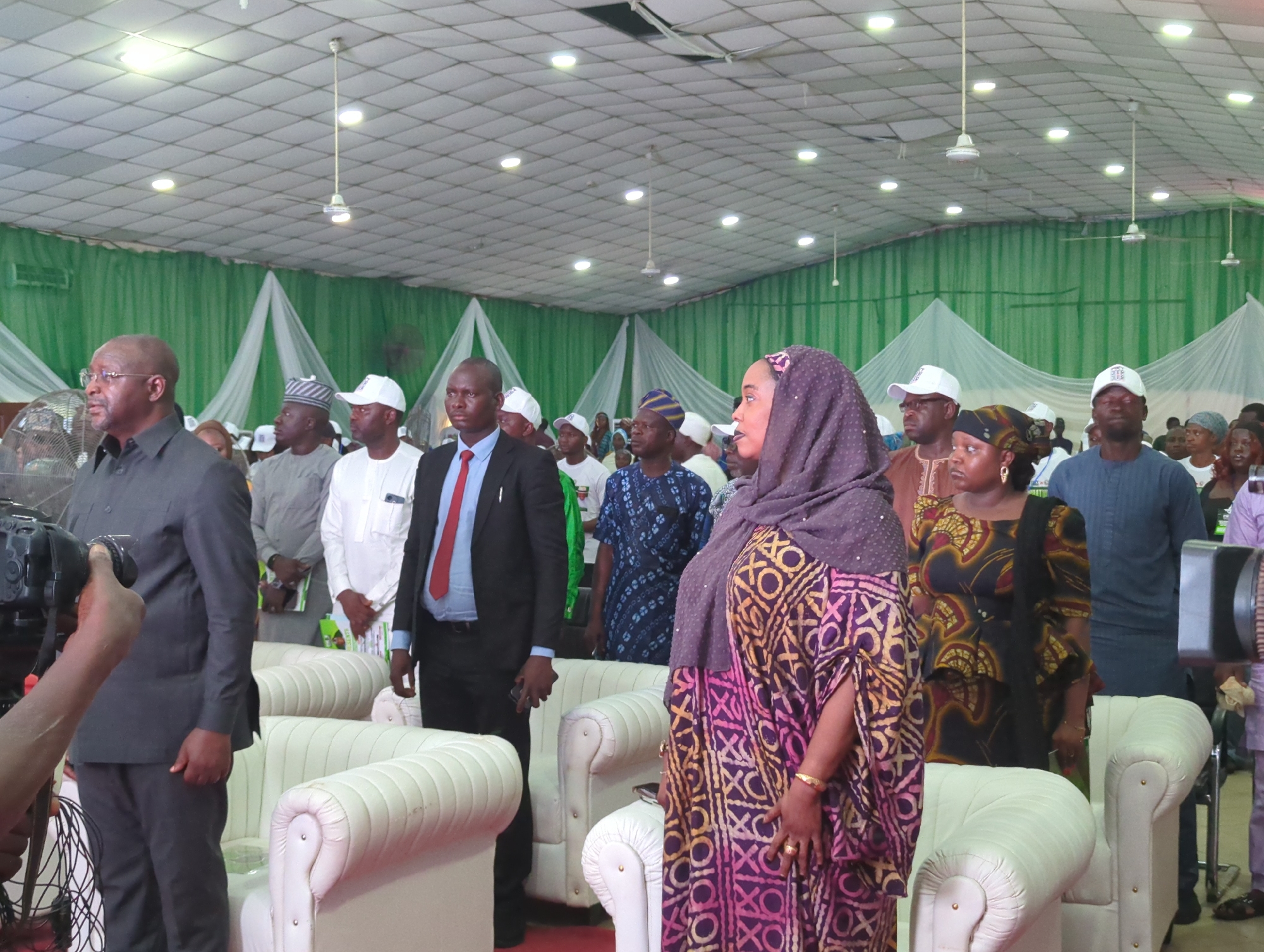 The image shows a group of people standing indoors, seemingly at an event. The location appears to be a large hall, indicated by the size, the drop ceiling with fluorescent lights, and several ceiling fans. Green and white draped fabric adorns the back of the room.
The people appear to be of Nigerian ethnicity. Several individuals are wearing traditional clothing.
A video camera is on the left side of the frame. There are white upholstered chairs lining the room.
The location is Ungwan Liman, Nigeria.