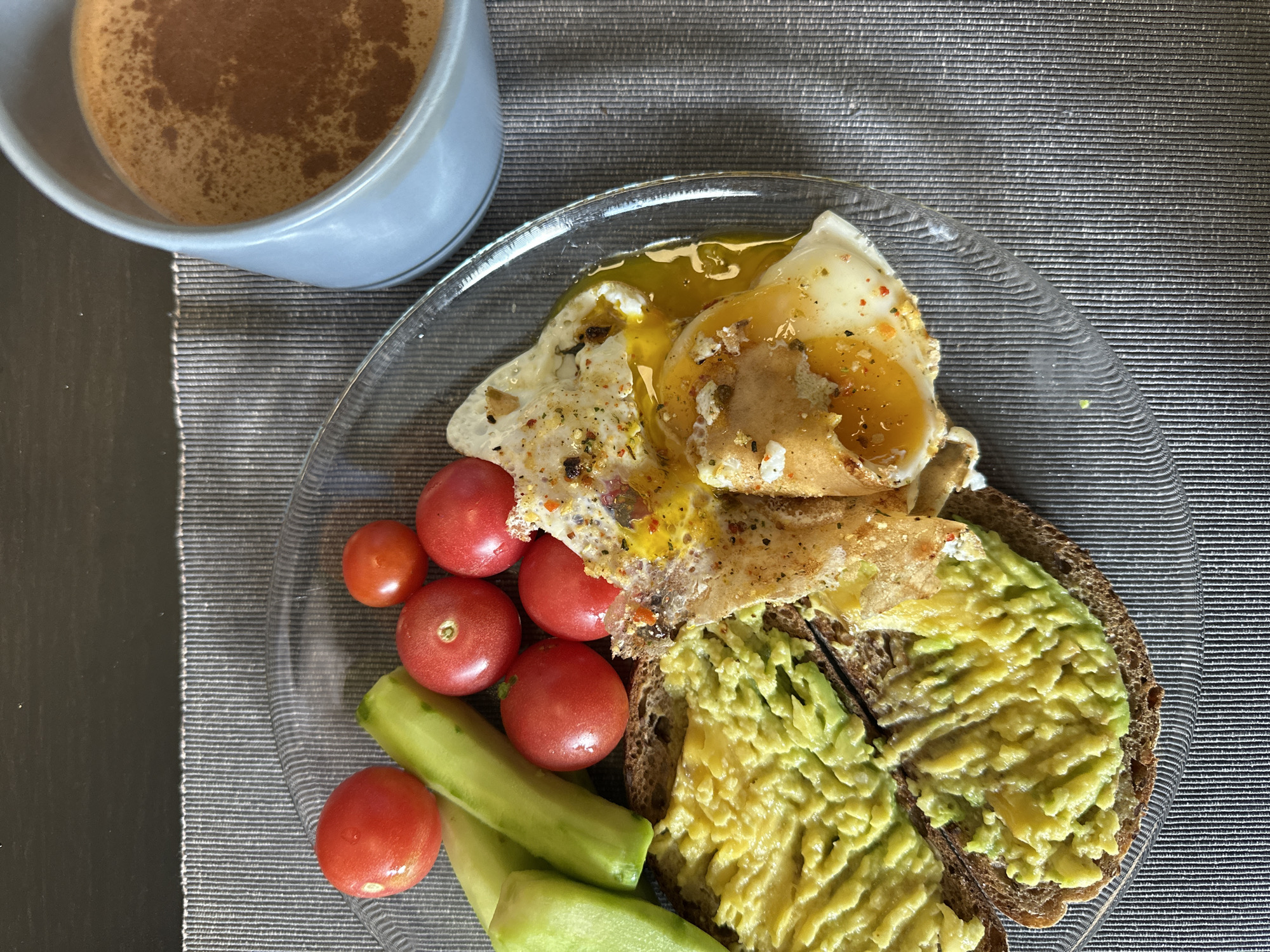 A close-up overhead shot of a breakfast plate with avocado toast, cherry tomatoes, cucumber slices, a fried egg, and a cup of coffee. The food is arranged on a clear glass plate, which sits on a grey textured placemat. The plate contains two slices of whole wheat toast, each generously spread with mashed avocado. To the left of the toast are several bright red cherry tomatoes and three diagonal slices of cucumber. A fried egg, with a runny yolk, is positioned on top of one of the toast slices. The egg is seasoned with a sprinkle of red pepper flakes and other spices. To the right of the plate, a light blue mug is filled with a frothy coffee beverage. The background is a dark, textured surface, possibly a table, with a hint of a darker grey area at the top. The lighting is natural and casts soft shadows. The scene is set in a home environment in Дачное, Russia.