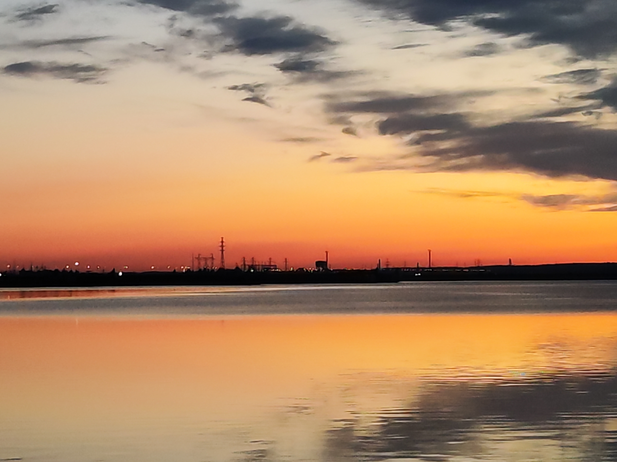 The image shows a body of water with reflections of the sky at sunset. The sky transitions from dark clouds above to an orange and red glow near the horizon. The dark silhouette of the shoreline and industrial structures is visible on the horizon. The location is likely near the city of Chaikovsky, Russia, given the presence of industrial structures and a large body of water which are typical for the Kama River area. No people are visible.