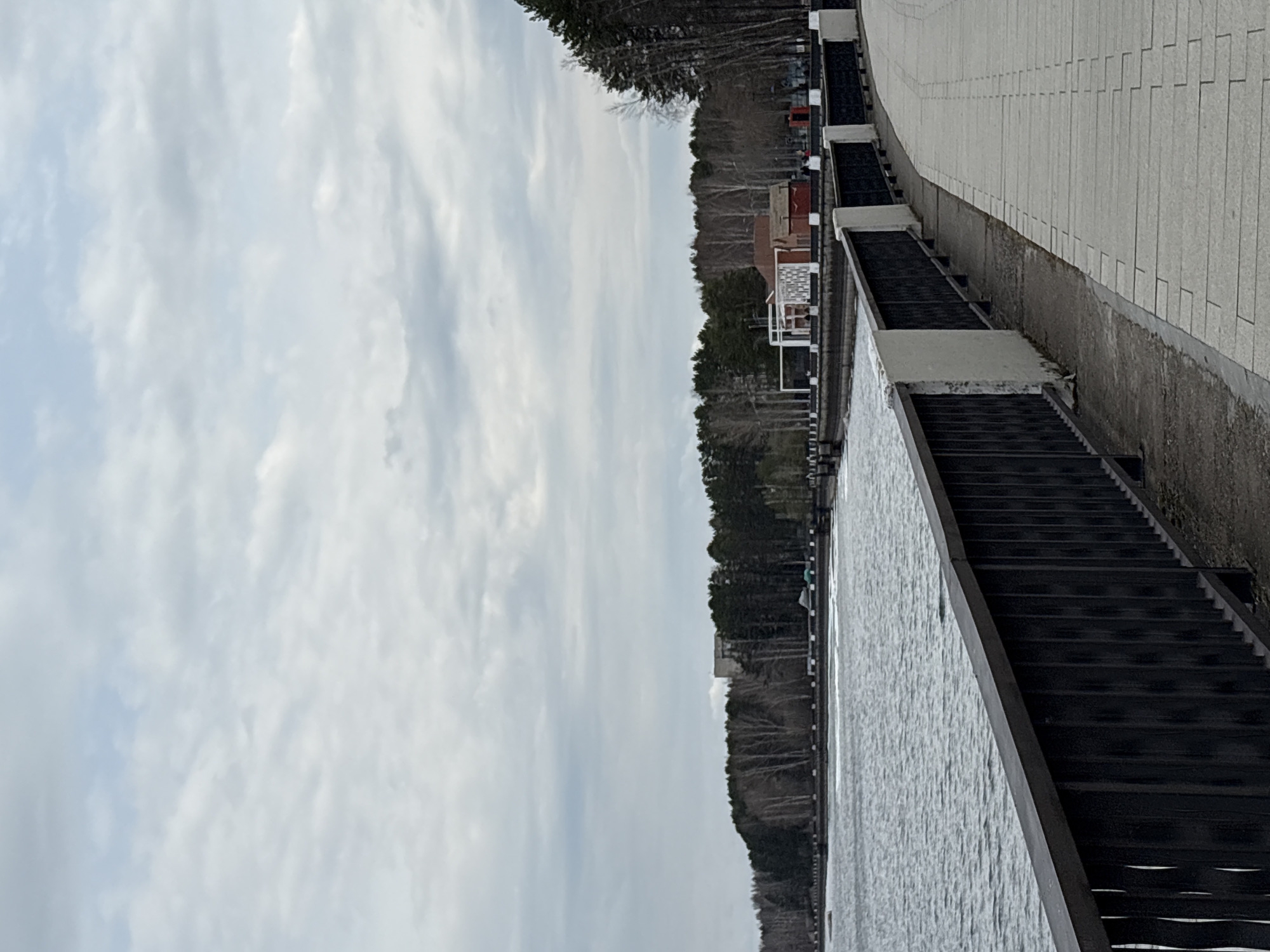 The image shows a stairway in Chaikovsky, Russia. The stairway descends towards a body of water (likely the Kama River). To the right of the stairway is a paved walkway. In the background, there are trees and a few buildings. The sky is cloudy.