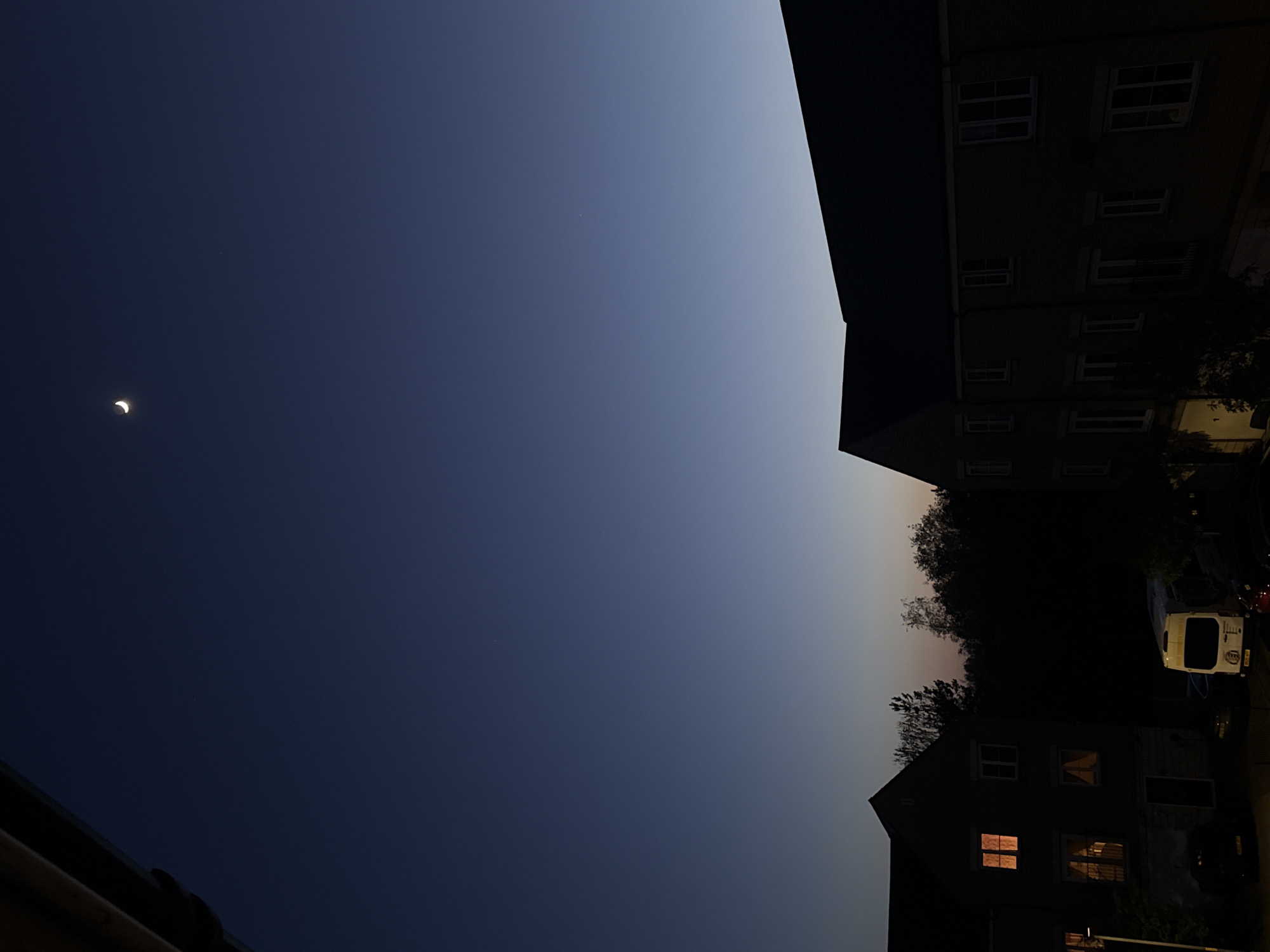 The image captures a serene twilight scene over a residential area, likely in Great Yarmouth, United Kingdom. The left side of the frame is dominated by a clear sky, displaying a beautiful gradient from a deep indigo at the far left to a lighter blue-gray and hints of orange-pink towards the right, indicating either dusk or dawn. A distinct, slender crescent moon is prominently visible in the darker, upper-left part of this sky. A very faint, small light, possibly a star or distant planet, can be discerned closer to the middle of the sky portion.
Along the right side and bottom right of the image, residential buildings and trees are silhouetted against the fading light. A dark, multi-story building frames the scene on the right. In the bottom right corner, a house with several warmly illuminated windows suggests interior lights are on. Trees are visible behind this house, silhouetted against the lighter part of the sky. A white van and a dark car are parked on what appears to be a street or driveway near the house. A dark roofline is also partially visible in the bottom left corner. The weather appears clear, with no clouds. No people or explicit activities are depicted, contributing to a calm and peaceful atmosphere. There is no visible text within the image.