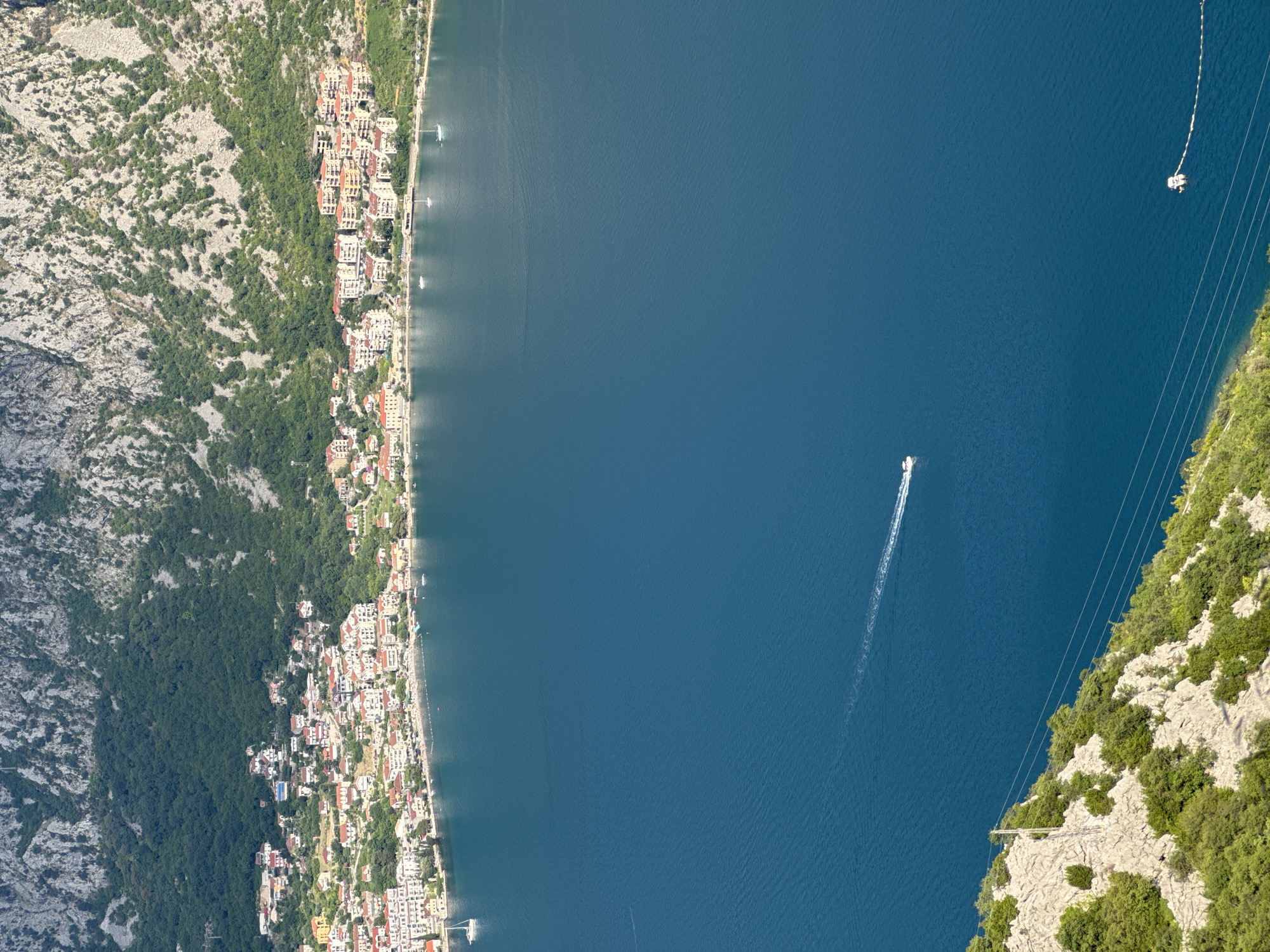The image captures a high-angle view of the Bay of Kotor, specifically looking down on the town of Štrp, Montenegro. A dense cluster of white and red-roofed buildings forms the coastal settlement, nestled at the base of a steep, rocky mountain covered in lush green vegetation. The deep blue water of the bay dominates the foreground and mid-ground. A white speedboat is seen creating a white wake as it travels across the water. Further out, a small white buoy is visible. On the left side of the image, a rocky, green-covered incline descends towards the water, with power lines running diagonally across it. The scene appears to be bathed in bright sunlight, casting shadows on the mountain.