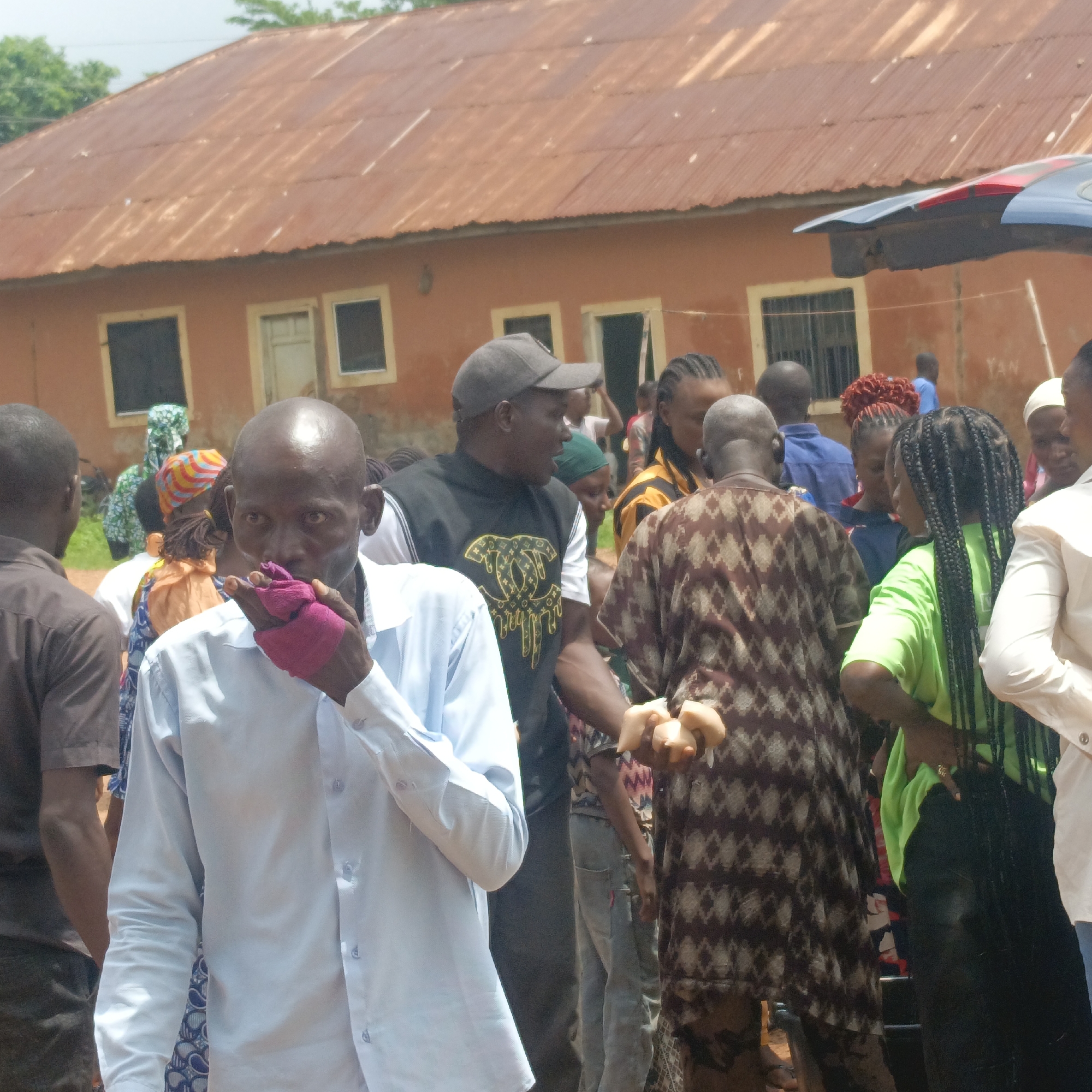 The image shows a group of people gathered outdoors in Akwanga, Nigeria. The foreground features a bald man in a light blue shirt holding a pink cloth to his mouth. Behind him, there is a man wearing a black baseball cap and a black t-shirt with a gold design. To the right, another man is holding several round, light-colored objects. Several other people are visible, some wearing bright colors or patterned clothing. A building with a rusty brown corrugated metal roof and dark-framed windows is visible in the background. A car roof can be seen on the right side of the building.