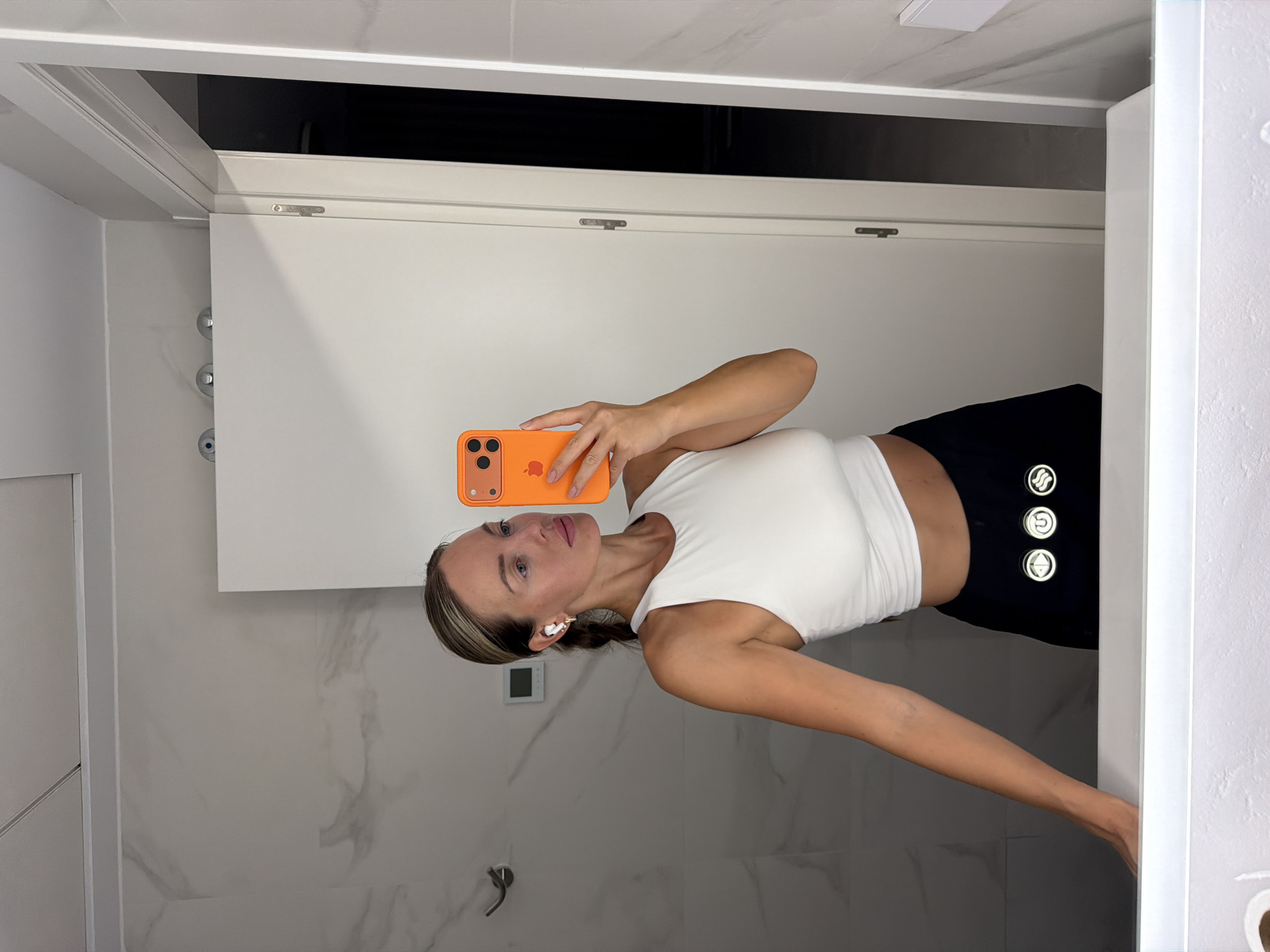 In Finestrat, Spain, a woman with blonde hair pulled back in a ponytail stands in a brightly lit bathroom, taking a selfie in a mirror. She is wearing a white crop top and black leggings with three illuminated circular logos on the left thigh. She has white earbuds in her ears and is holding an orange smartphone. The bathroom features white marble walls, a white vanity with a bright light underneath, and a white door.