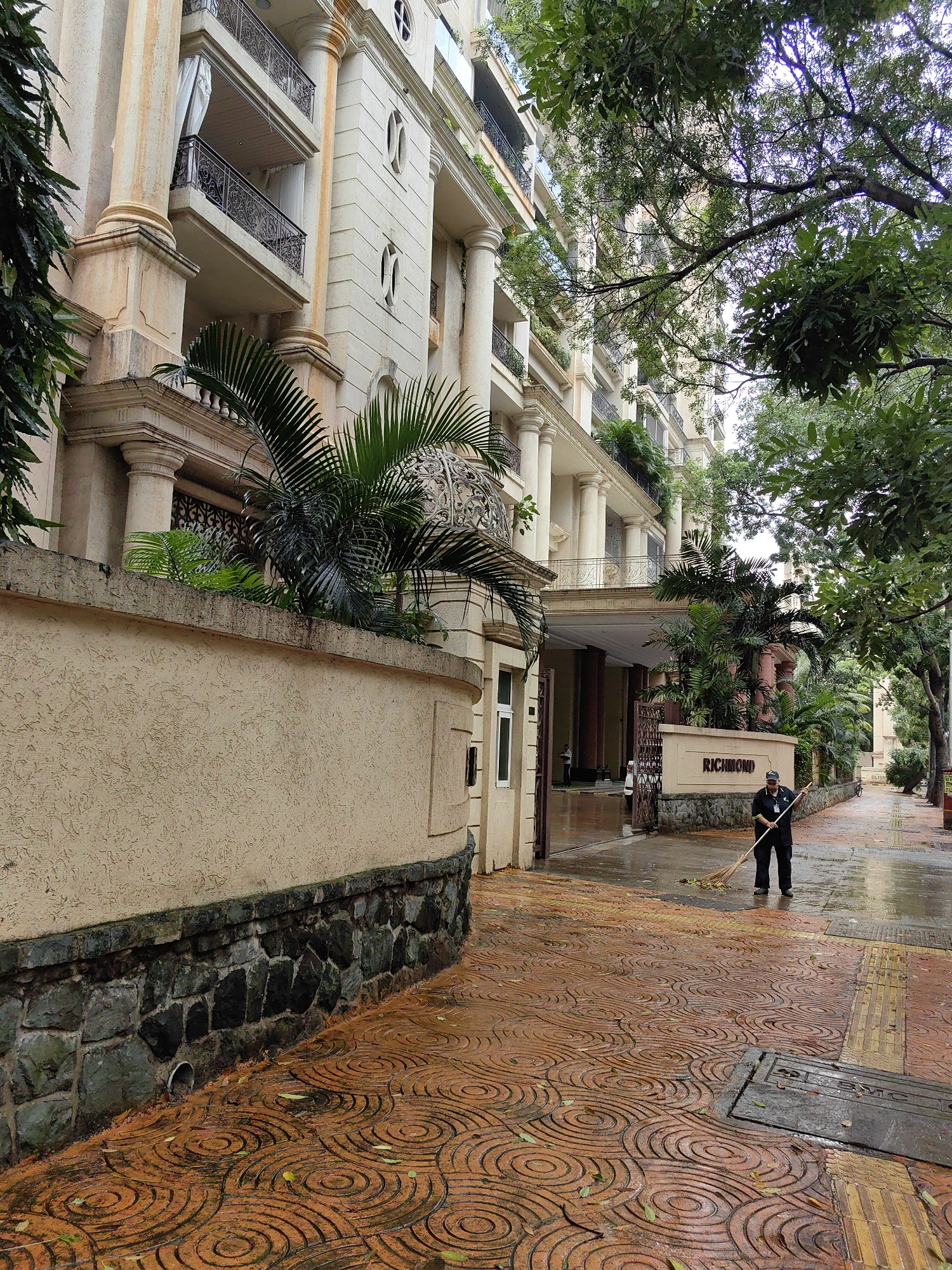 An exterior shot of the Richmond building in Mumbai, India, depicts a man sweeping leaves on a wet sidewalk. The building features a light-colored facade with tall columns and balconies, partially obscured by lush green trees. The sidewalk is paved with circular-patterned reddish-brown tiles, some of which are wet, suggesting recent rainfall. A stone wall with a textured surface separates the sidewalk from the building's entrance, which is marked by a sign that reads "RICHMOND." The man, dressed in a dark uniform, is actively sweeping leaves towards a pile with a broom. The overall atmosphere is one of an urban environment experiencing a light shower, with a focus on the building's architecture and the activity of the groundskeeper.