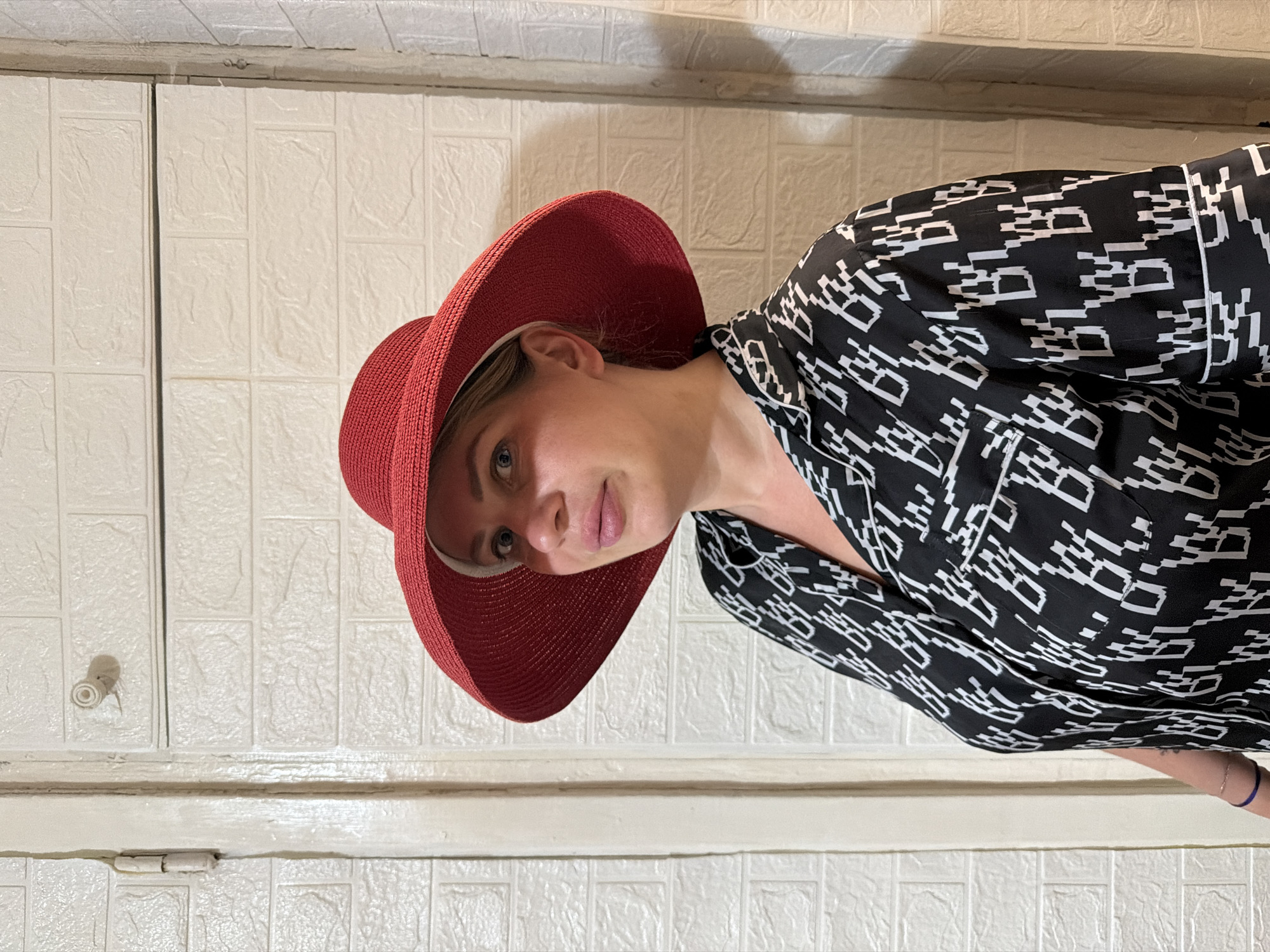 A Caucasian woman with blonde hair and blue eyes is posing for a photo indoors, likely in Sochi, Russia. She is wearing a red wide-brimmed hat and a black pajama-style top with a repeating white pattern. The background consists of a textured white wall that resembles brickwork, and a white doorframe with a hinge visible on the left side. A small, round object resembling a knob or a fixture is attached to the wall. The lighting suggests an indoor setting.