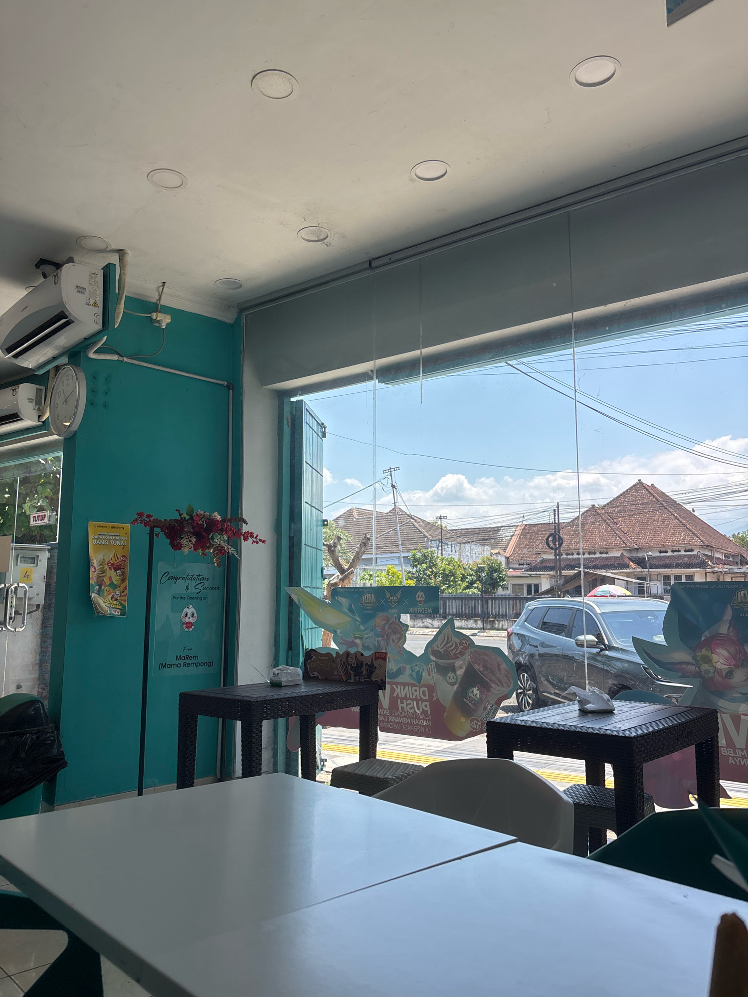 The image captures an interior view of a shop in Jember, Indonesia, looking out through a large glass window. The interior features a turquoise wall with an air conditioning unit mounted high on it, a clock, and a poster advertising a product or service, with text in Indonesian. A bouquet of red flowers is placed on a stand next to a sign that reads "Congratulations & Success For the opening of" and "From MaRem (Mama Rempong)".
Inside, several dark wicker tables and chairs are arranged. One table has a white napkin dispenser on it.
Through the glass window, a street scene in Jember is visible. Buildings with terracotta roofs dominate the background, with power lines and lush green trees interspersed. A dark-colored SUV is parked on the street, and various posters or cutouts with cartoon-like characters are displayed in front of the shop window, some with text in Indonesian. The sky is blue with scattered white clouds. The overall impression is that of a casual establishment, possibly a cafe or a retail shop, in a warm, sunny climate.