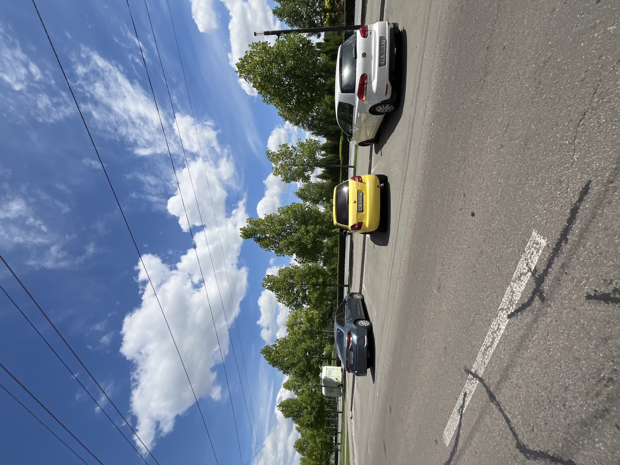 The image shows a street scene with three cars parked in a row. The cars are a white Volkswagen Golf, a yellow compact car, and a dark gray sedan. The cars are parked on an asphalt road with white painted lines marking parking spots. Trees line the side of the road, and in the background, there is a blue sky with white, puffy clouds. Power lines are visible diagonally across the sky. The sun appears to be high in the sky, suggesting it is daytime. The weather appears to be clear and sunny. The license plates of the cars are partially visible: the white car has a plate starting with "AE 0600 ET", and the yellow car has a plate starting with "AE 0772 ET". The dark gray car has a license plate that is difficult to read but appears to start with "CZ". The scene is a typical street in an urban or suburban area. The image is oriented vertically. The location is specified as Dnipro, Ukraine.
