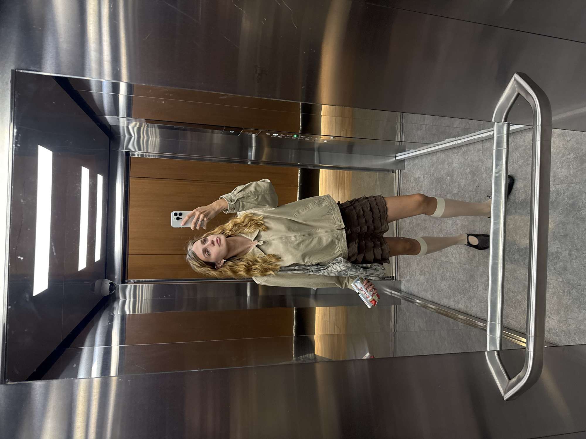A female is photographed inside an elevator. She is holding a smartphone in her right hand, presumably taking a selfie. She is wearing a light-colored, button-up jacket, a short brown skirt, white socks, and black shoes. In her left hand, she holds a package of what appears to be a drink. The elevator has stainless steel walls, a wooden panel, and a handrail. The floor appears to be gray tile. The location and city are not identifiable from the image.
