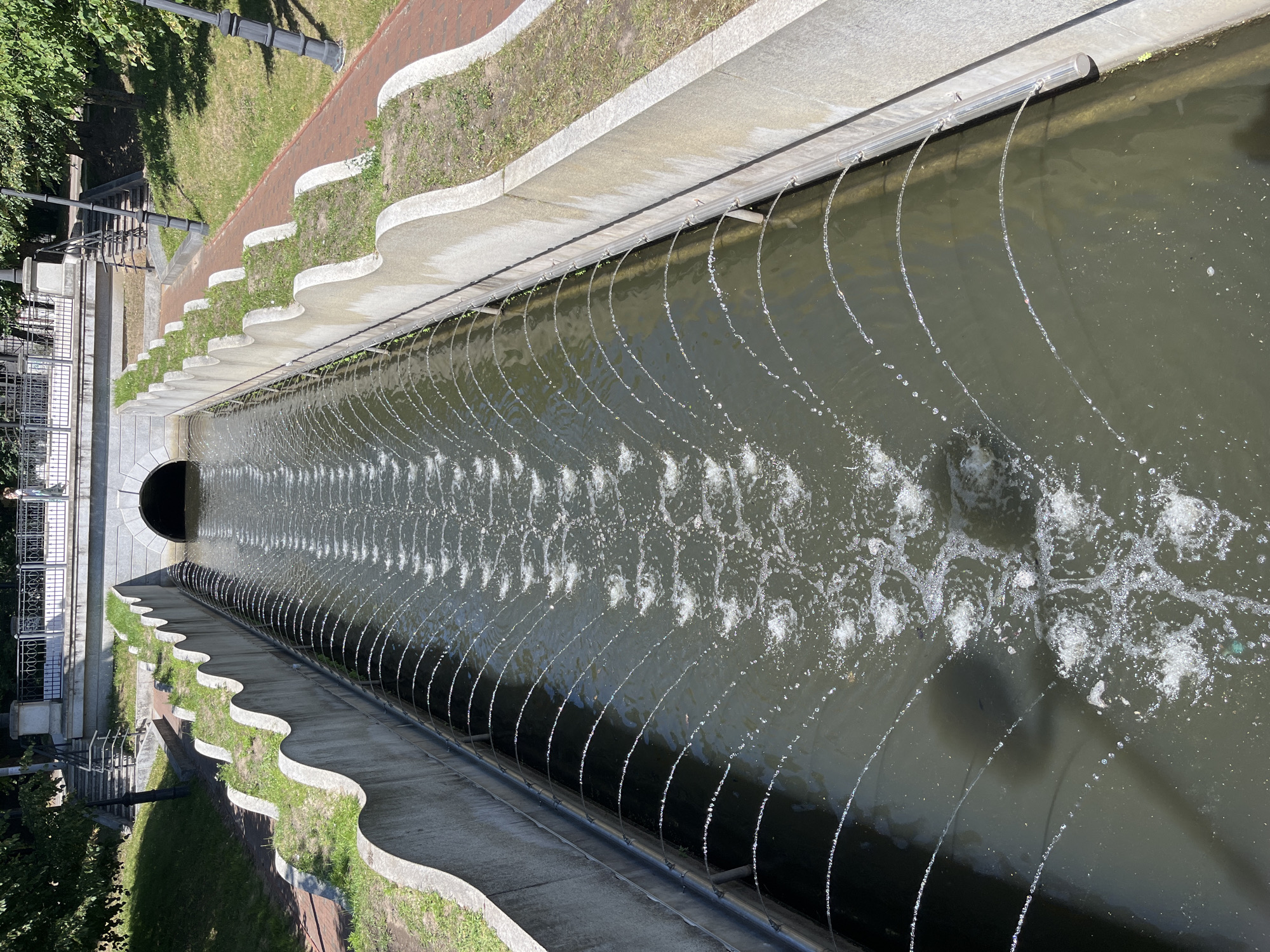 The image shows a water channel with fountains spraying water in arcs on both sides. The channel is lined with concrete walls that have a wavy design on the upper edge, with grass growing in the indentations. To the right of the channel, there is a red brick pathway and a grassy area. On the left side of the channel, there are stairs leading up to a walkway with a railing, and trees in the background. A man is standing on the walkway in the distance. The channel appears to be leading towards a stone archway, suggesting it might be part of a larger waterway or park feature in Kaliningrad, Russia.