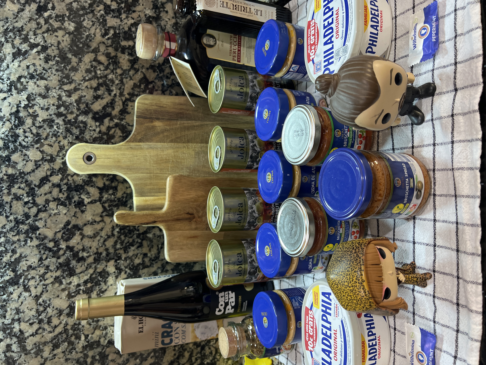 This media file captures a collection of food items, beverages, and decorative figures arranged on a kitchen countertop in Mollet Del Vallès, Spain. The setting features a mottled grey and black granite-patterned surface on the left, transitioning to a white and black checkered cloth on the right.
The scene presents various objects, including four wooden cutting boards of different sizes stacked centrally. Above the boards, a dark bottle of "VERMOUTH PERUCCHI GRAN RESERV" is visible, alongside "LE TRIBUTE TONIC WATER". Below the boards, another dark bottle, labeled "Con Par PREMIUM", rests on a box that mentions "EL HO PANA mini CRA COM S" (likely crackers), next to a small glass bottle with a cork stopper.
Vertically aligned are several canned goods, all from the "Jolca Artes de Maestros Aceituneros" brand, suggesting they contain olives. Adjacent to these are multiple glass jars, mostly with blue lids, some marked "CUCINA". Contents include "CREMA DE ATUN" (tuna spread) and "BRUSCHETTA SPREAD" in olive and cherry tomato flavors. One jar with a silver lid displays a date, "PB44/26A 28/02/28".
On the checkered cloth, several large tubs of "PHILADELPHIA ORIGINAL" cream cheese are visible, with text in Spanish stating "Queso delicioso y cremoso" and "ELABORADO EN ESPAÑA," along with promotional offers like "10€ Gratis En restaurantes" and "FORMATO FAMILIAR." Two Funko Pop! figures are placed among the items: one with dark hair in a bun and a black outfit, and another with blonde/leopard print hair and a leopard print outfit. A white wrapper for "Wilhelmina PEPERMUNT" is also present. The arrangement suggests a display of groceries or ingredients, possibly for a meal or gathering, with no direct activity taking place.