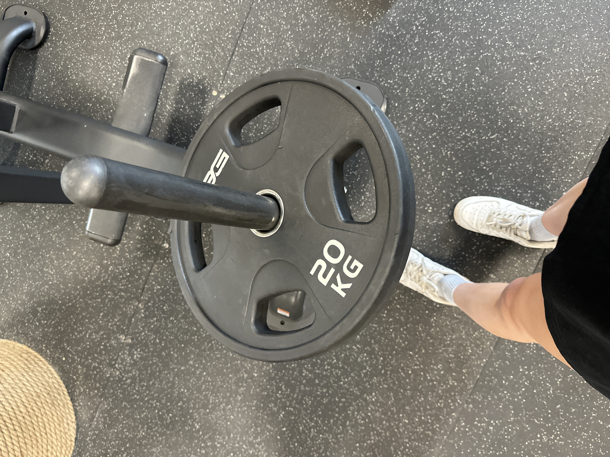 The image shows a 20 kg weight plate on a barbell. The weight plate is black with white lettering. The barbell is mounted on a black metal frame. The floor is a speckled gray. The lower legs and feet of a person wearing white sneakers and black athletic shorts are visible in the lower right of the frame. A round object, possibly a hat, is visible in the lower left of the frame. There is no information in the image to determine the location in the city of Yerevan, Armenia.