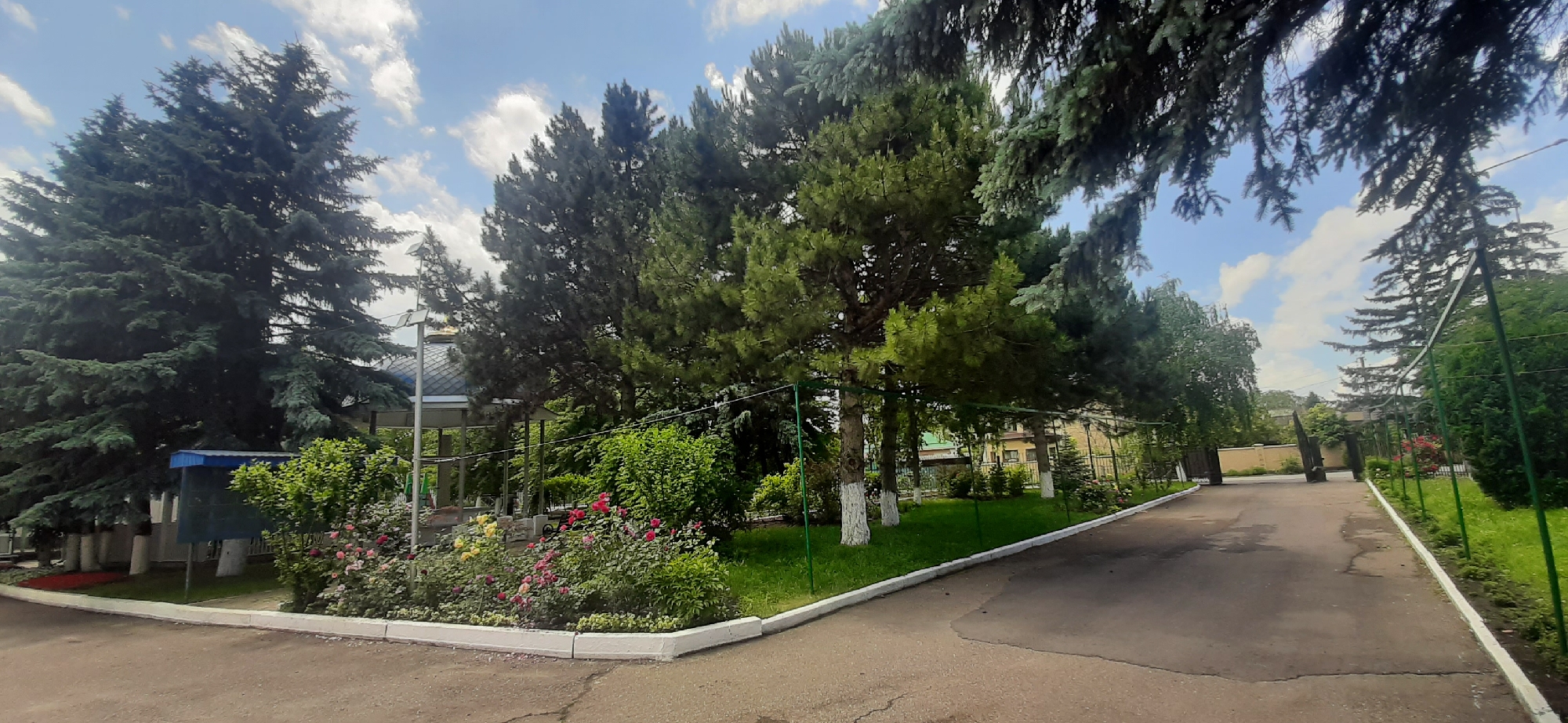 The image depicts a paved pathway leading through a garden area in Yessentukskaya, Russia. The pathway curves to the right and appears to lead towards an open gate. Lush greenery dominates the scene, with a variety of trees, including tall evergreen conifers and smaller deciduous trees, providing ample shade. A bed of colorful roses is visible in the foreground, adding a vibrant touch to the landscape. A small structure with a blue roof stands to the left, and a building with yellow siding is visible in the background. The sky is partly cloudy, with white clouds scattered across a blue expanse. There are no people or animals visible in the image.