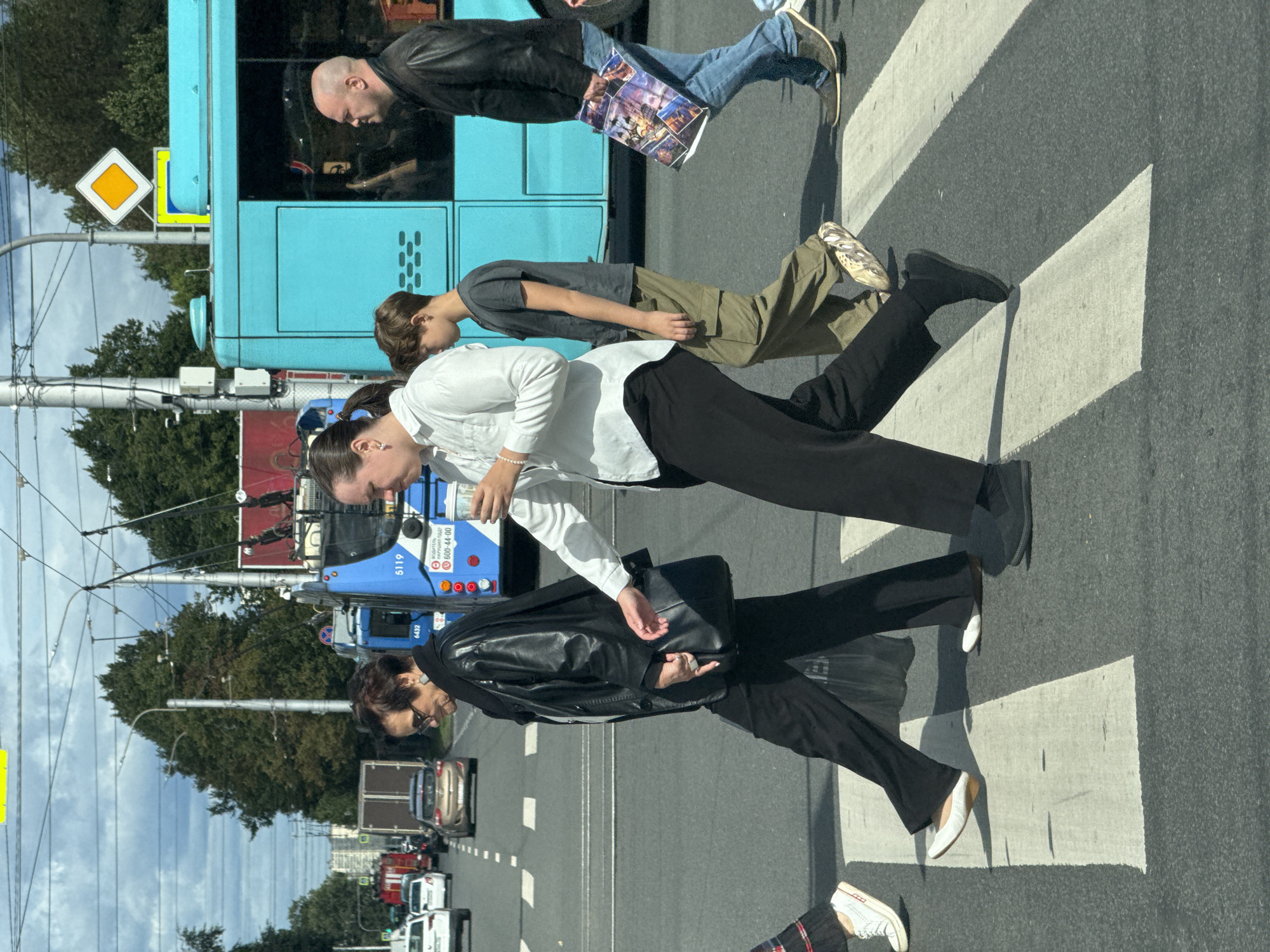 A group of pedestrians is crossing a street at a crosswalk. The scene takes place in the city of Ladozhskoe Ozero, Russia, with a bright blue trolleybus and other vehicles visible in the background.
In the foreground, an older woman with short dark hair, wearing a black leather jacket, black pants, and sunglasses, is walking to the left. She is carrying a black handbag. Next to her, a young woman with her hair pulled back in a ponytail, wearing a white shirt and black pants, is walking to the left, holding a white cup. Behind them, a young boy in a grey t-shirt and green cargo pants is walking to the left. To the far right, a bald man wearing a black jacket and blue jeans is walking to the left, holding a folded magazine. Another person's legs are visible on the far left, wearing white sneakers. The pedestrians are on a marked crosswalk, indicated by white stripes on the asphalt. Overhead power lines for the trolleybus are visible, along with trees and buildings in the distance. The sky is cloudy.