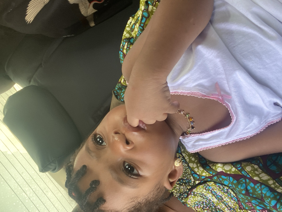 A young child is lying down wearing a white shirt with pink trim and a colorful beaded necklace. One hand is near the child's mouth. The child has dark hair styled in braids and is wearing a small earring. The child is lying on a colorful patterned fabric. Part of a black couch is visible in the background.