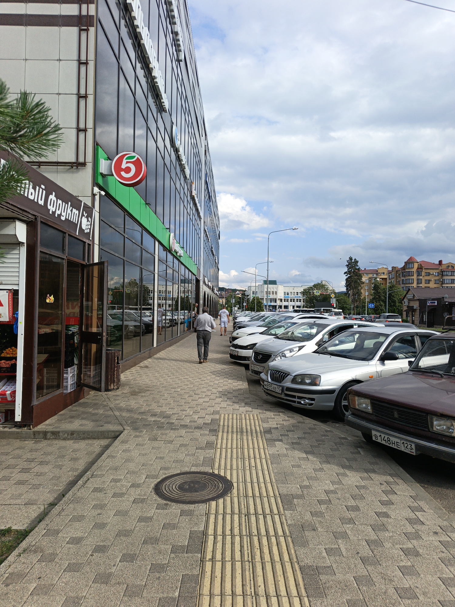 The image depicts a street scene in Goryachiy Klyuch, Russia, with a large modern building featuring a prominent "5" logo on its facade. The building has a glass exterior and appears to be a commercial establishment. In front of the building, a sidewalk paved with interlocking concrete blocks runs parallel to a row of parked cars. A man in a light-colored shirt and dark pants is walking away from the camera on the sidewalk. A small shop with an open door and displays of fruit is visible to the left of the building. The sky is partly cloudy, and trees and other buildings are visible in the background. The parked cars include several silver sedans and a darker colored vehicle in the foreground.