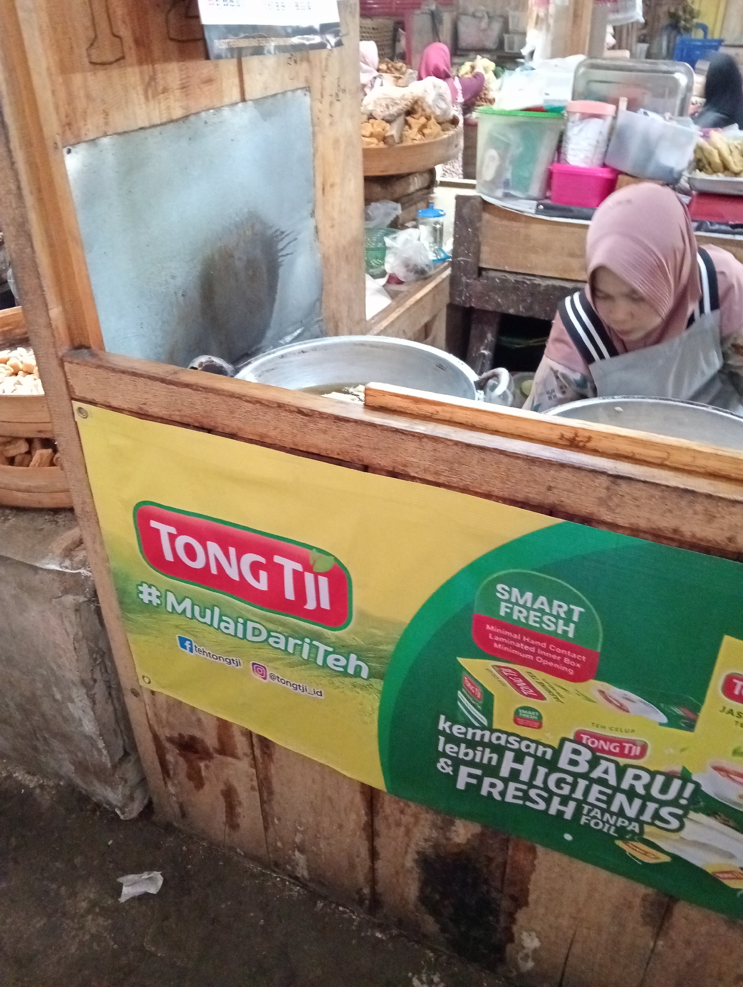 The image depicts a market scene in Grabag, Indonesia. In the foreground, a yellow banner with red lettering reads "TONG TJII" and "#MulaiDariTeh". Below this, social media icons are visible with associated handles. A woman wearing a pink hijab and an apron is partially visible in the background, attending to food preparation. Various food items and containers are scattered throughout the scene, suggesting a bustling marketplace environment. Wooden structures and concrete surfaces indicate the typical setting of an Indonesian market.