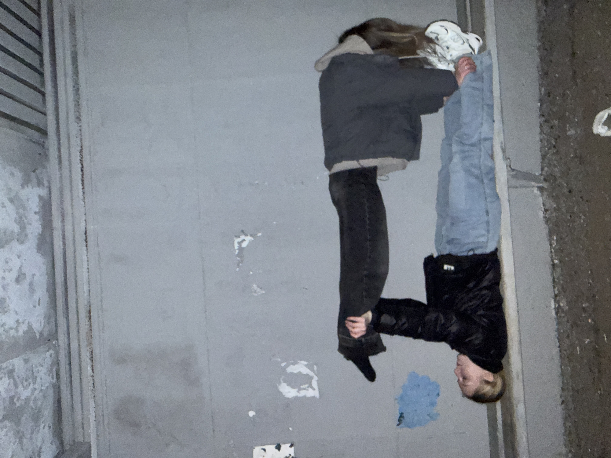 Two individuals are juxtaposed against a nondescript wall in an urban setting, potentially located in an area with a Russian cultural influence based on the query origin, although no definitive regional indicators are visible in the image. One person is oriented with their head upwards, wearing white sneakers, light blue jeans, and a dark jacket. They appear to be holding the ankles of the other individual. The second person is inverted, wearing a gray jacket, dark jeans, and black shoes. The wall behind them is gray and features patches of discoloration and minor graffiti. The overall setting suggests an alleyway or a similar utilitarian urban space.